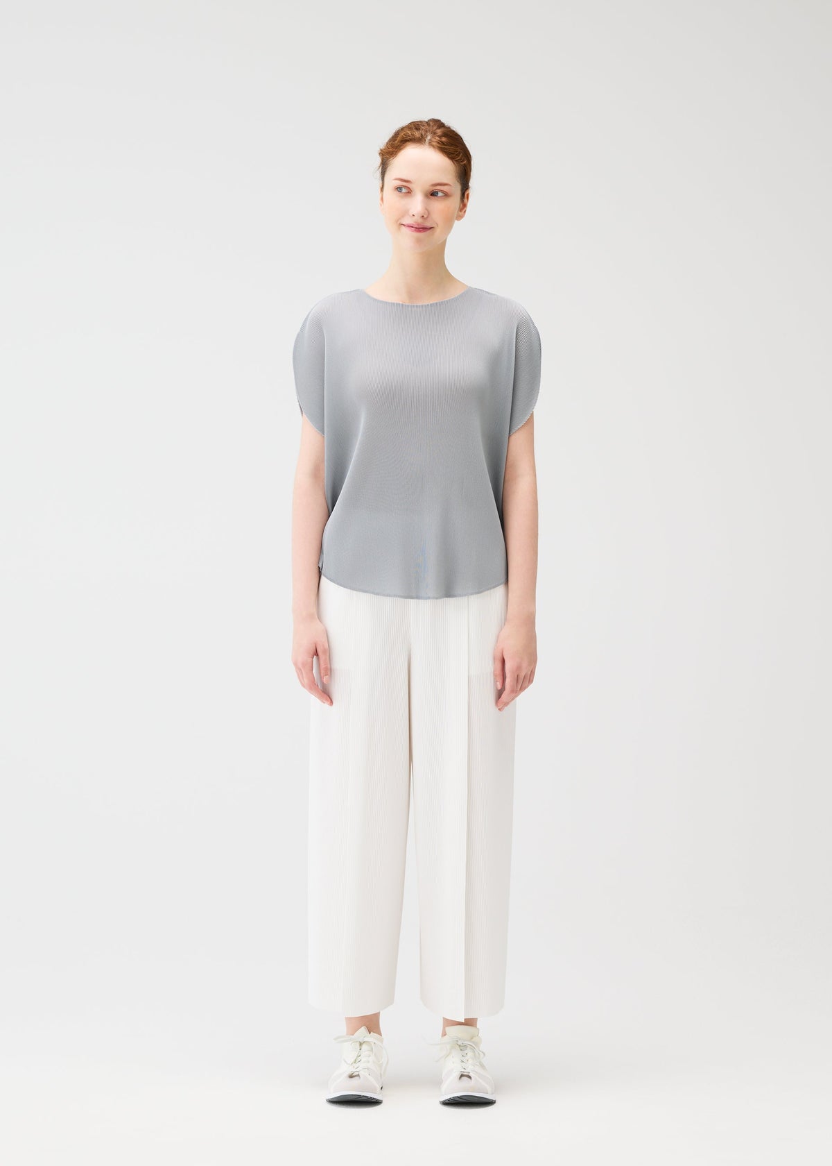 A woman with pulled-back hair and a slight smile stands facing forward against a plain white background, wearing the light gray MIST APRIL pleated top by PLEATS PLEASE ISSEY MIYAKE, wide white pants, and white sneakers. view 6 of 8