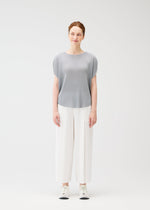 A woman with pulled-back hair and a slight smile stands facing forward against a plain white background, wearing the light gray MIST APRIL pleated top by PLEATS PLEASE ISSEY MIYAKE, wide white pants, and white sneakers.