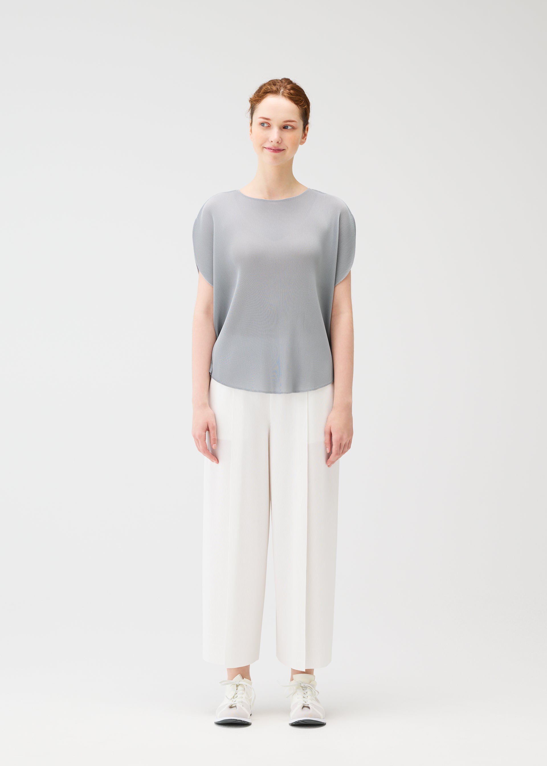 Zoomed view A woman with pulled-back hair and a slight smile stands facing forward against a plain white background, wearing the light gray MIST APRIL pleated top by PLEATS PLEASE ISSEY MIYAKE, wide white pants, and white sneakers.