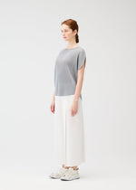 A woman stands in a minimal studio wearing the gray MIST APRIL circle top by PLEATS PLEASE ISSEY MIYAKE, paired with wide white pants and white sneakers. She faces left with a neutral expression against a plain white background.