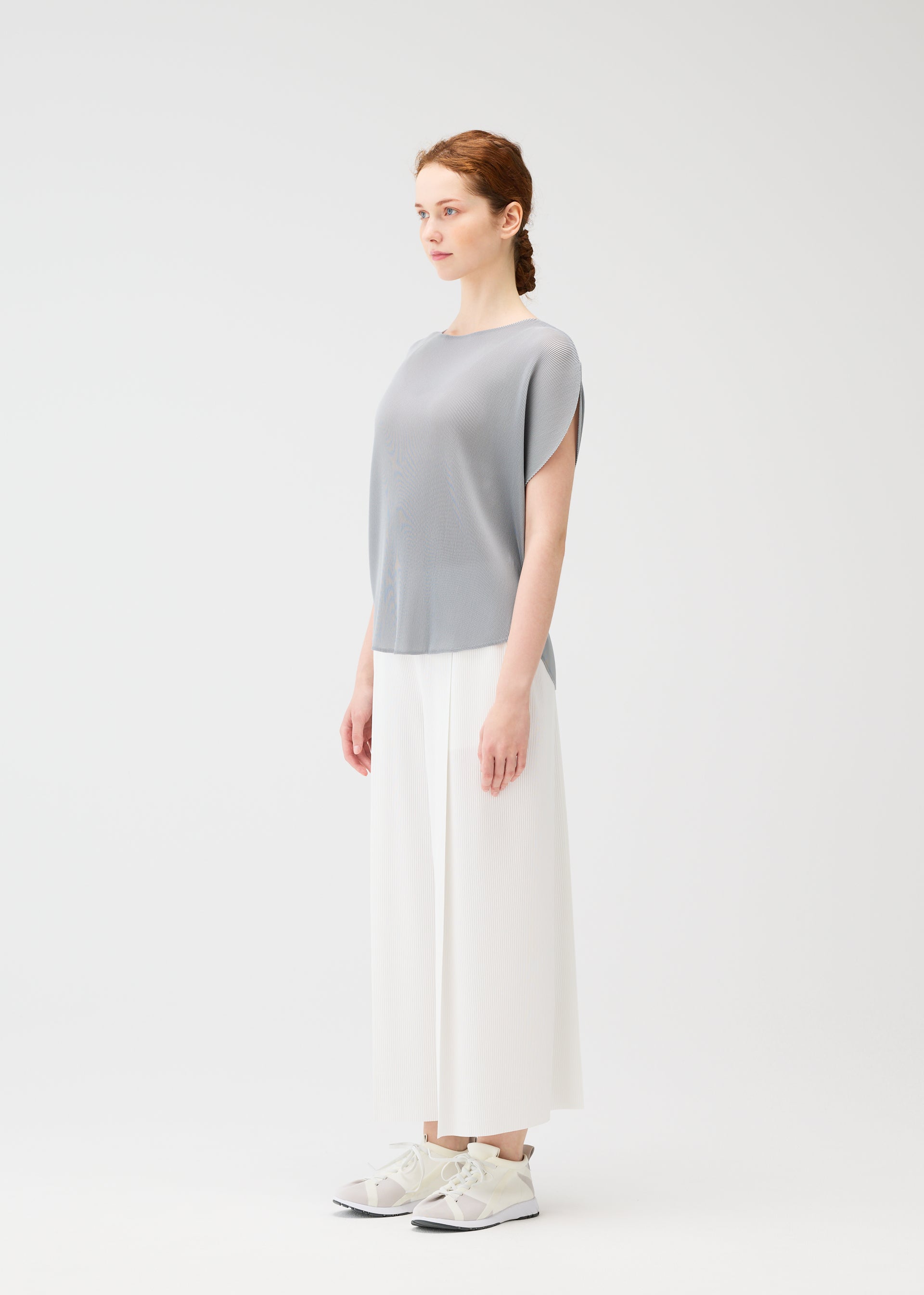 Zoomed view A woman stands in a minimal studio wearing the gray MIST APRIL circle top by PLEATS PLEASE ISSEY MIYAKE, paired with wide white pants and white sneakers. She faces left with a neutral expression against a plain white background.