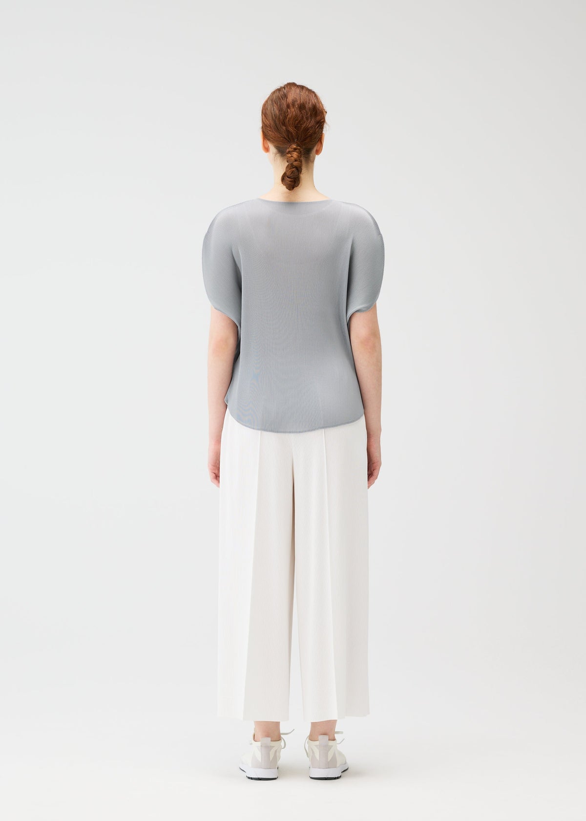 A person with red hair in a low bun faces away, wearing the MIST APRIL top by PLEATS PLEASE ISSEY MIYAKE, wide white pants, and white sneakers against a plain white background. view 8 of 8