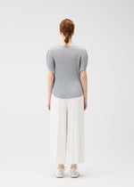 A person with red hair in a low bun faces away, wearing the MIST APRIL top by PLEATS PLEASE ISSEY MIYAKE, wide white pants, and white sneakers against a plain white background.