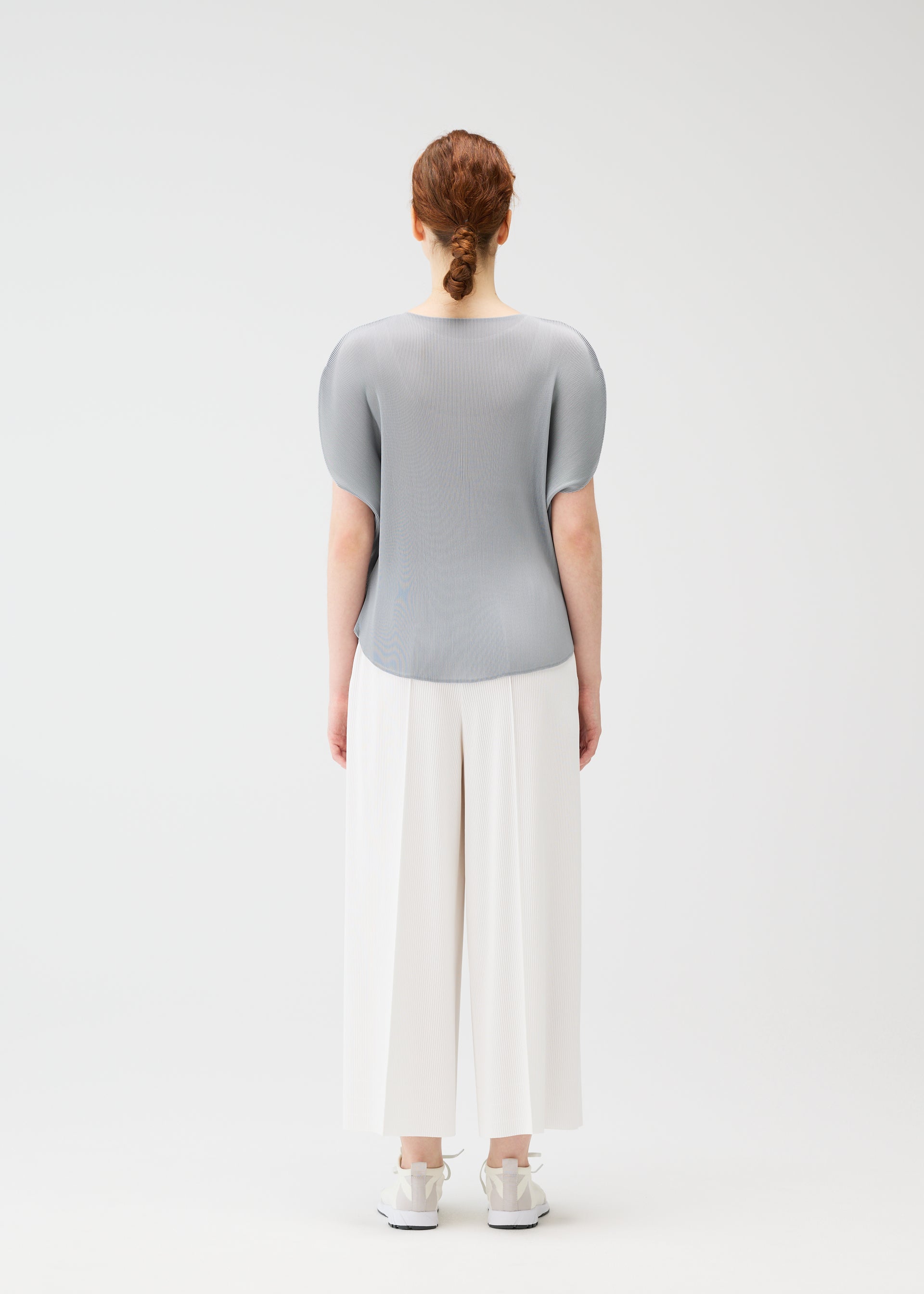 Zoomed view A person with red hair in a low bun faces away, wearing the MIST APRIL top by PLEATS PLEASE ISSEY MIYAKE, wide white pants, and white sneakers against a plain white background.