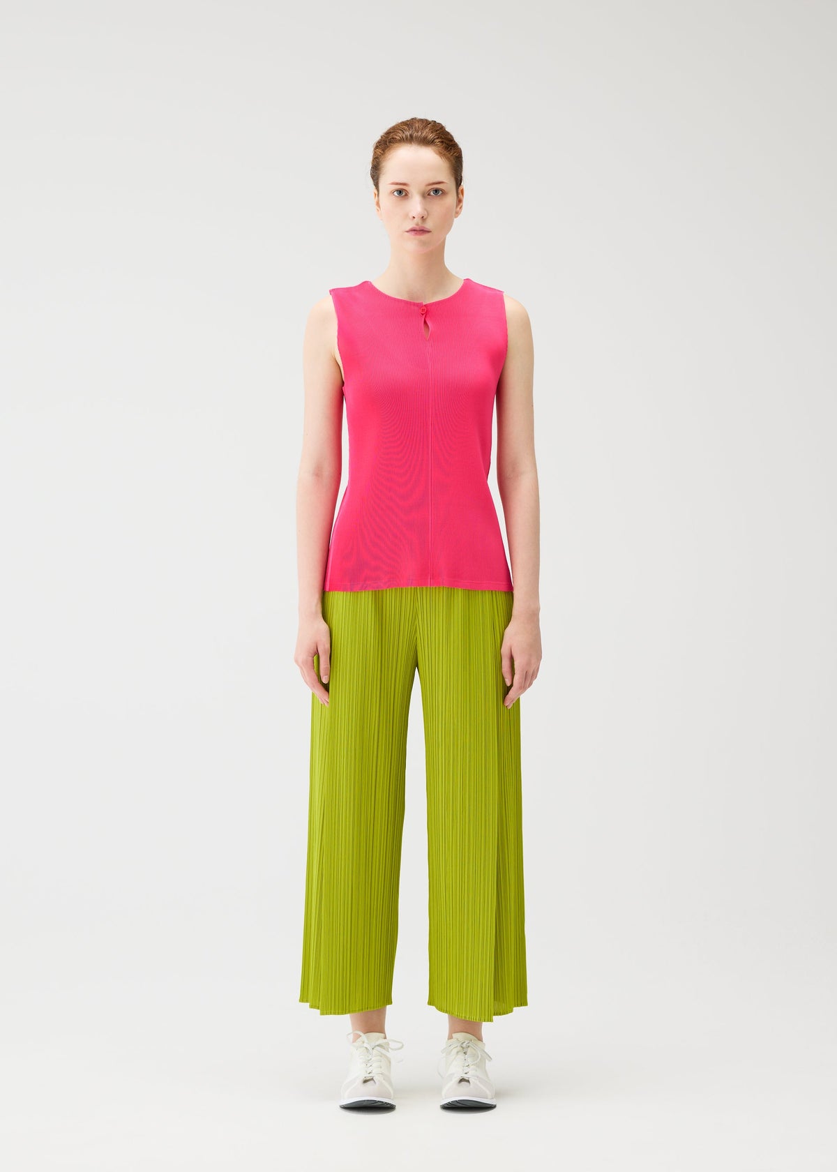 A woman faces forward against a white background, wearing the MIST JUNE bright pink tank top and lime green pleated wide-leg pants by PLEATS PLEASE ISSEY MIYAKE, styled with white sneakers and a neutral expression. view 5 of 7
