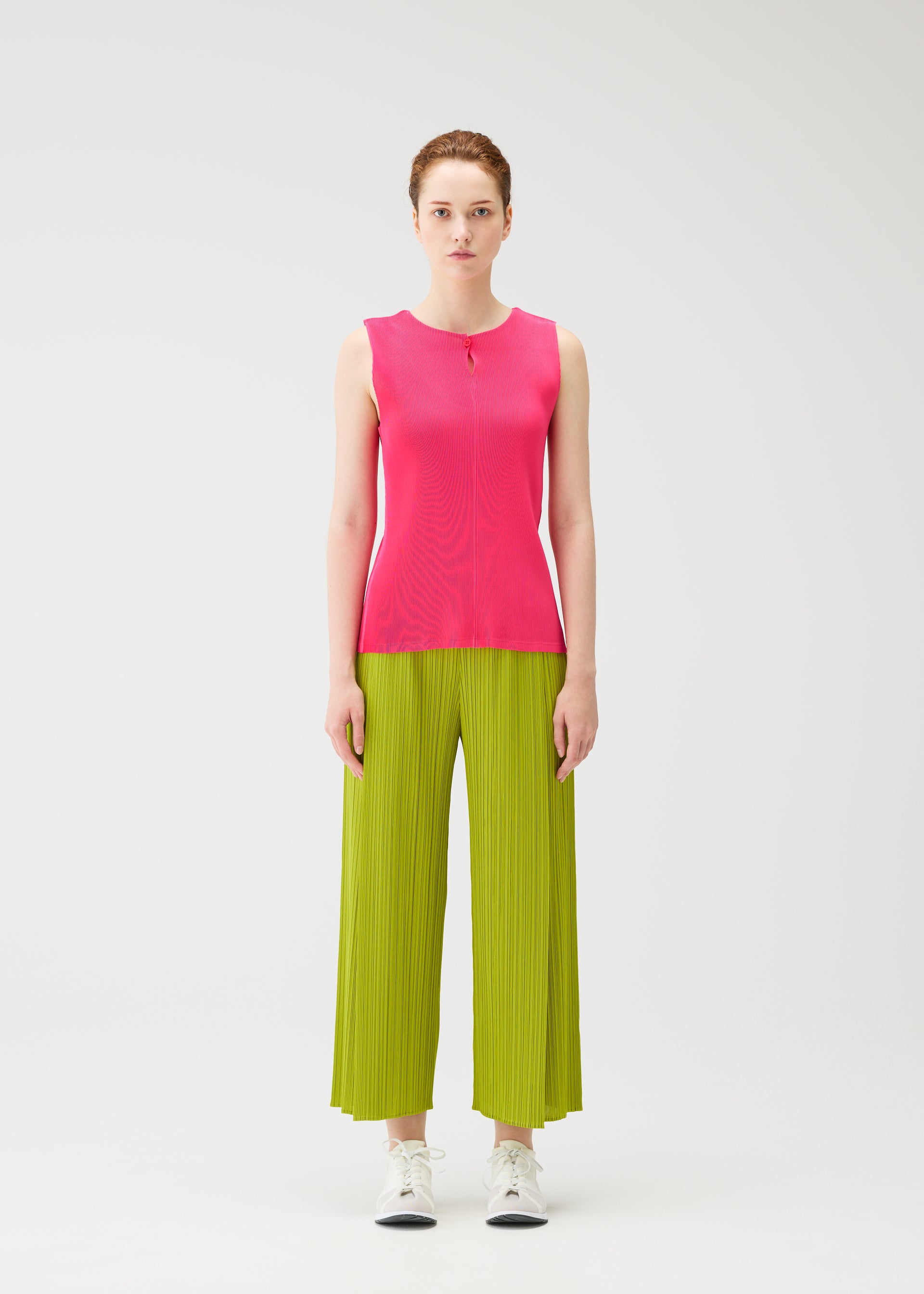 Zoomed view A woman faces forward against a white background, wearing the MIST JUNE bright pink tank top and lime green pleated wide-leg pants by PLEATS PLEASE ISSEY MIYAKE, styled with white sneakers and a neutral expression.