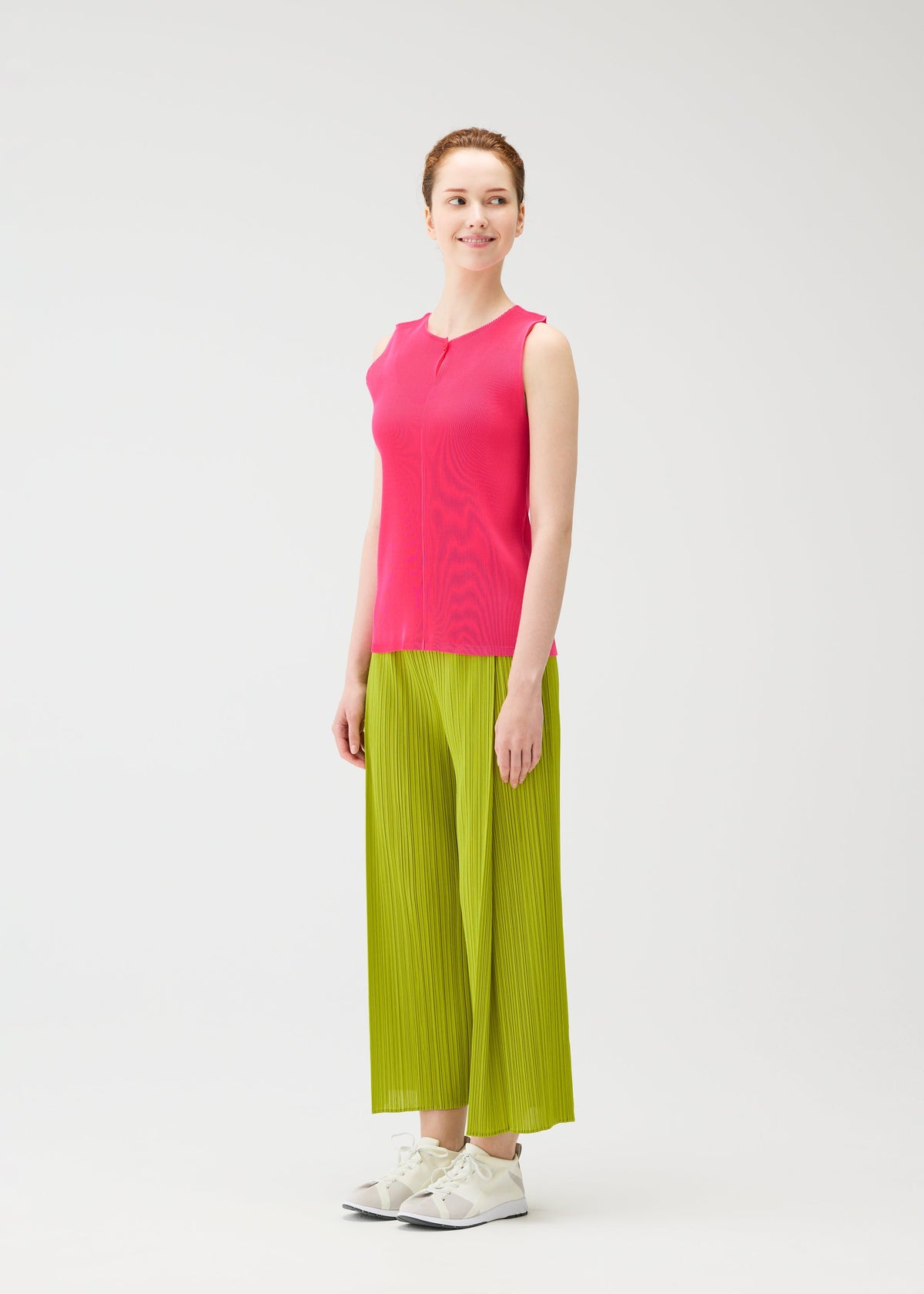 A person smiles in MIST JUNE by PLEATS PLEASE ISSEY MIYAKE: a bright pink lightweight tank top and wide-leg lime green pants, styled with white sneakers, posed against a plain white background. view 6 of 7