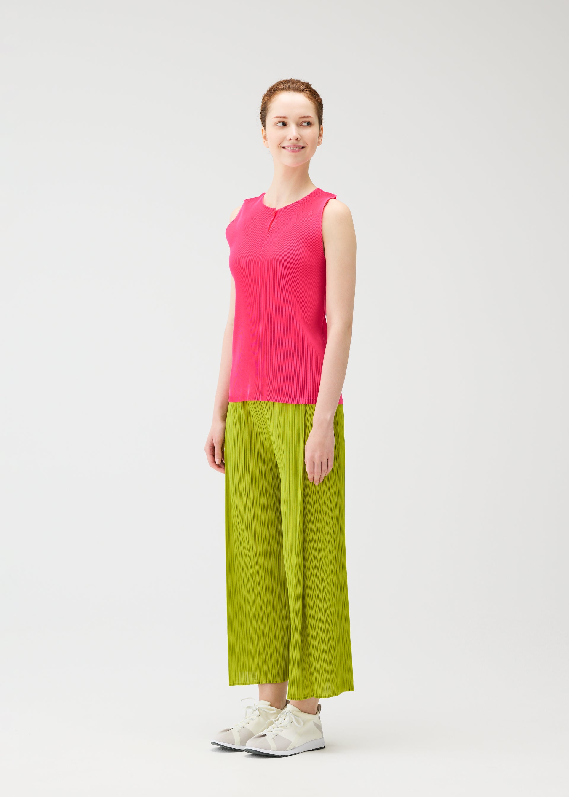 Zoomed view A person smiles in MIST JUNE by PLEATS PLEASE ISSEY MIYAKE: a bright pink lightweight tank top and wide-leg lime green pants, styled with white sneakers, posed against a plain white background.