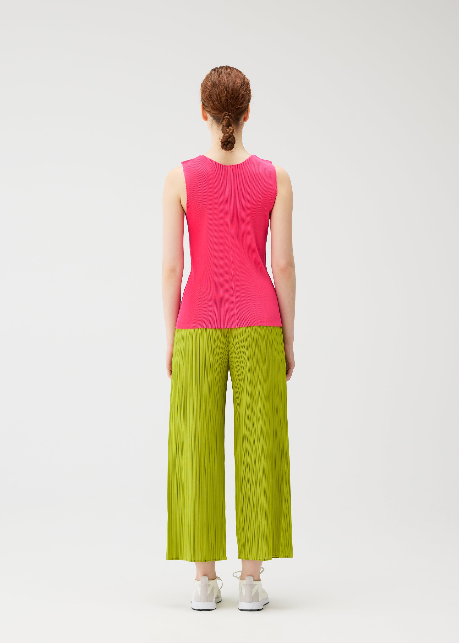 Zoomed view A person with tied-back hair faces away, wearing the MIST JUNE sleeveless bright pink top and lime green pleated wide-leg pants by PLEATS PLEASE ISSEY MIYAKE, paired with white sneakers against a plain white background.