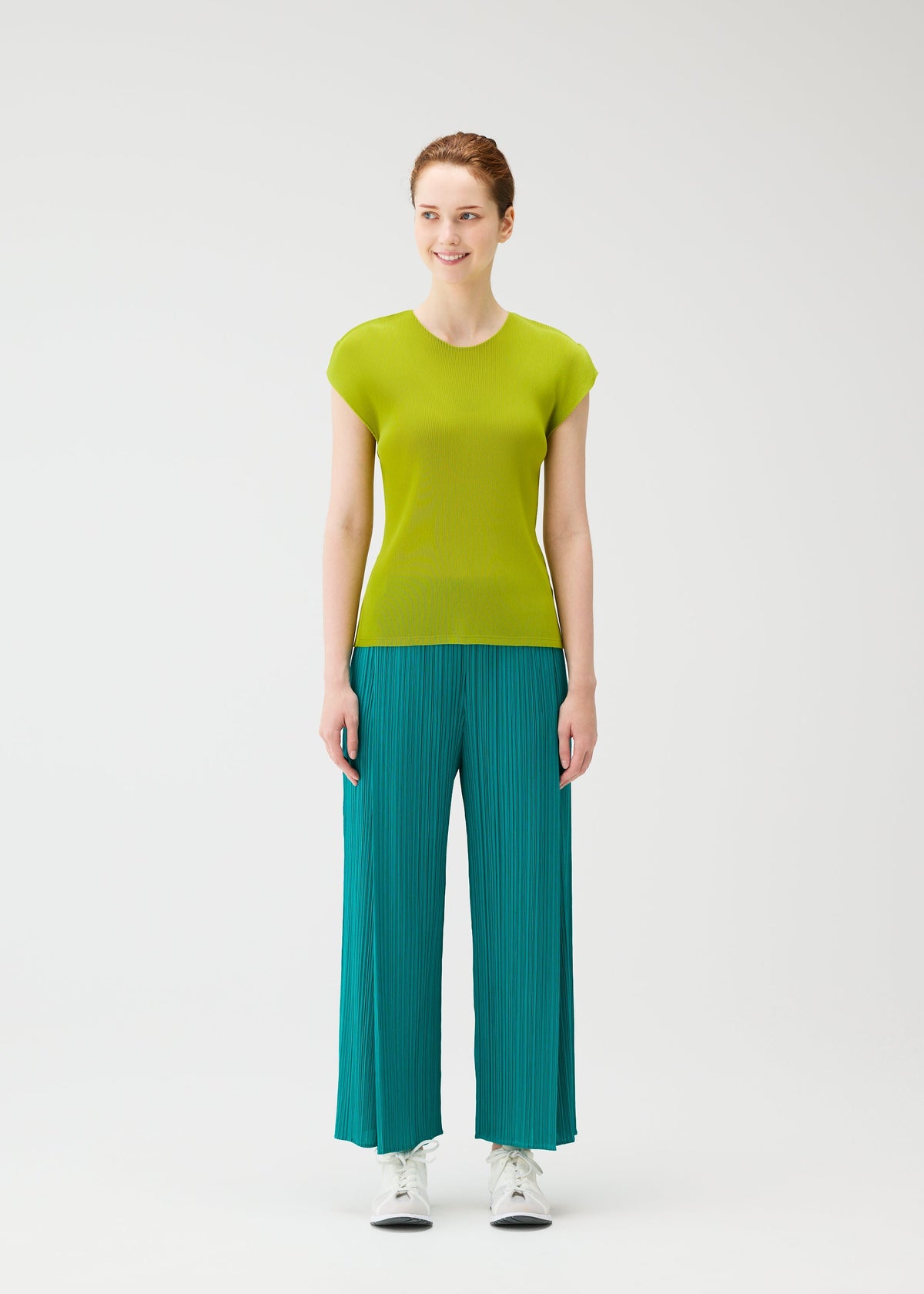 A woman stands against a white background, smiling and looking aside. She wears the MIST JUNE top and fine-pleated loose teal pants by PLEATS PLEASE ISSEY MIYAKE, paired with crisp white sneakers. view 5 of 7