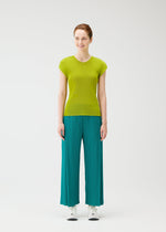 A woman stands against a white background, smiling and looking aside. She wears the MIST JUNE top and fine-pleated loose teal pants by PLEATS PLEASE ISSEY MIYAKE, paired with crisp white sneakers.