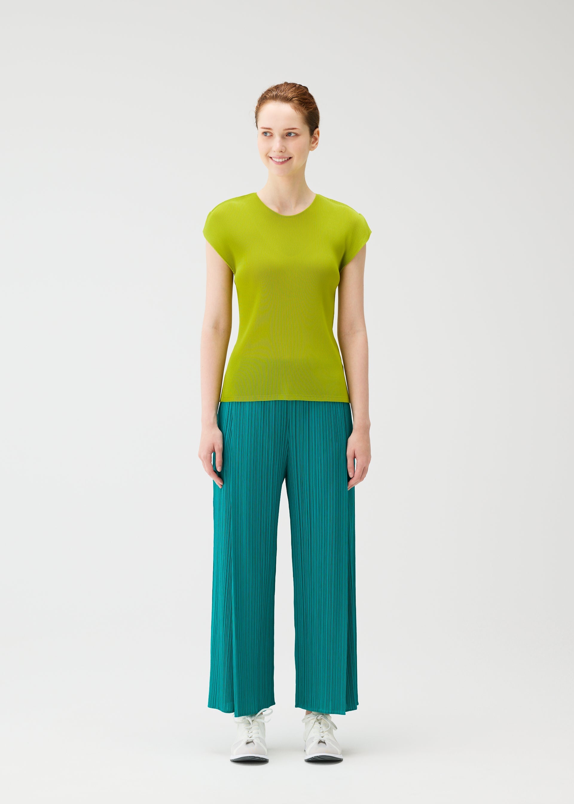 Zoomed view A woman stands against a white background, smiling and looking aside. She wears the MIST JUNE top and fine-pleated loose teal pants by PLEATS PLEASE ISSEY MIYAKE, paired with crisp white sneakers.