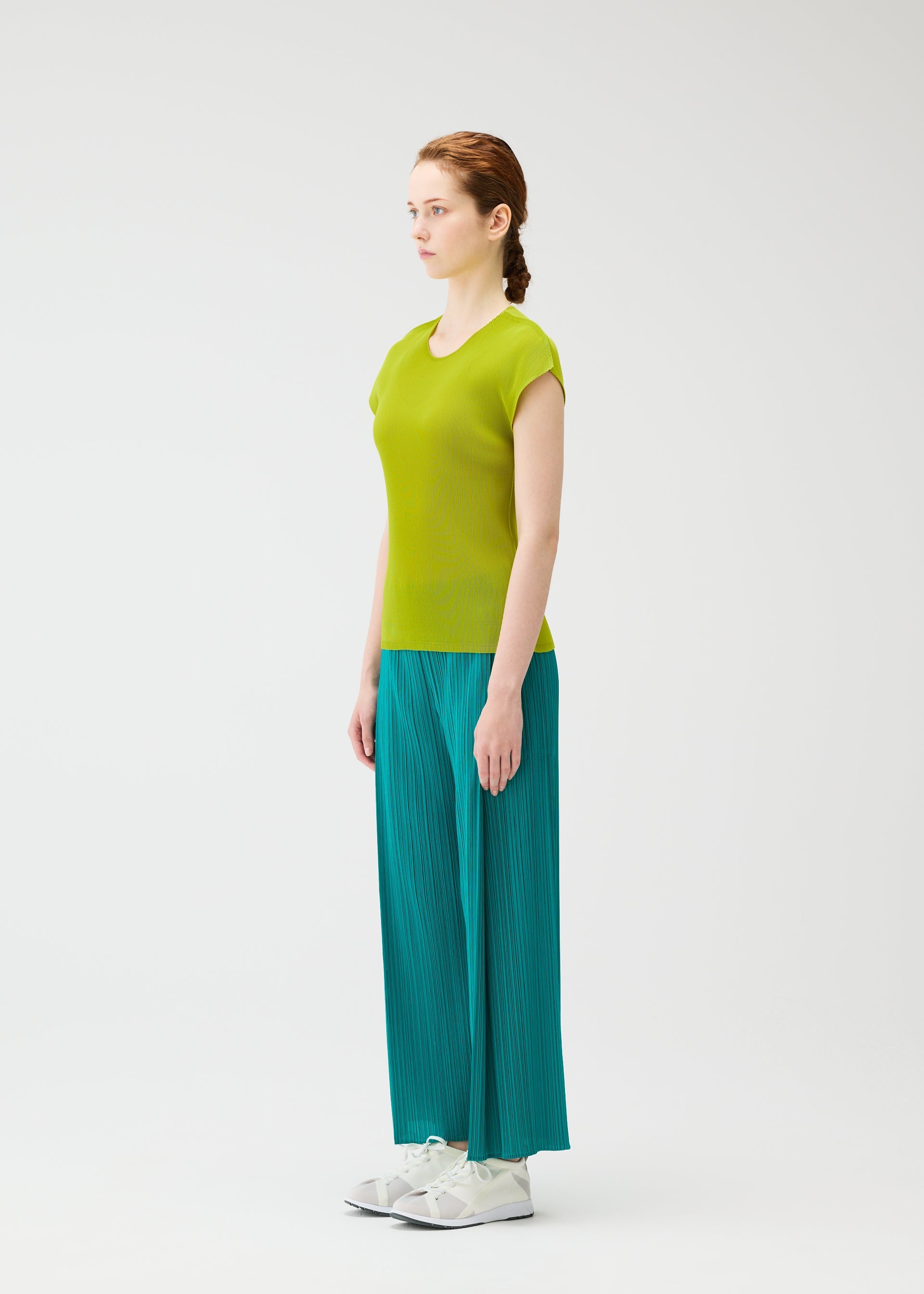 Zoomed view A woman in profile stands against a white background, wearing the MIST JUNE top from PLEATS PLEASE ISSEY MIYAKE in lime green with fine pleats, paired with teal wide-leg pants and white sneakers, her hair in a neat braid.