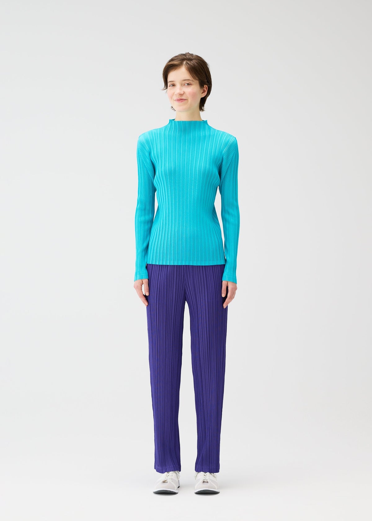 A person with short brown hair and a neutral expression stands facing forward against a white background, wearing the PLEATS PLEASE ISSEY MIYAKE RIB PLEATS JANUARY turquoise top and dark blue pleated pants with white shoes. view 5 of 7