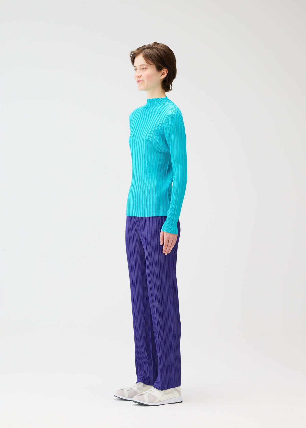 A person stands in profile against a plain white background, wearing PLEATS PLEASE ISSEY MIYAKE’s RIB PLEATS JANUARY sweater with loose-fitting purple pants and white shoes. view 6 of 7