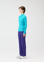 A person stands in profile against a plain white background, wearing PLEATS PLEASE ISSEY MIYAKE’s RIB PLEATS JANUARY sweater with loose-fitting purple pants and white shoes.