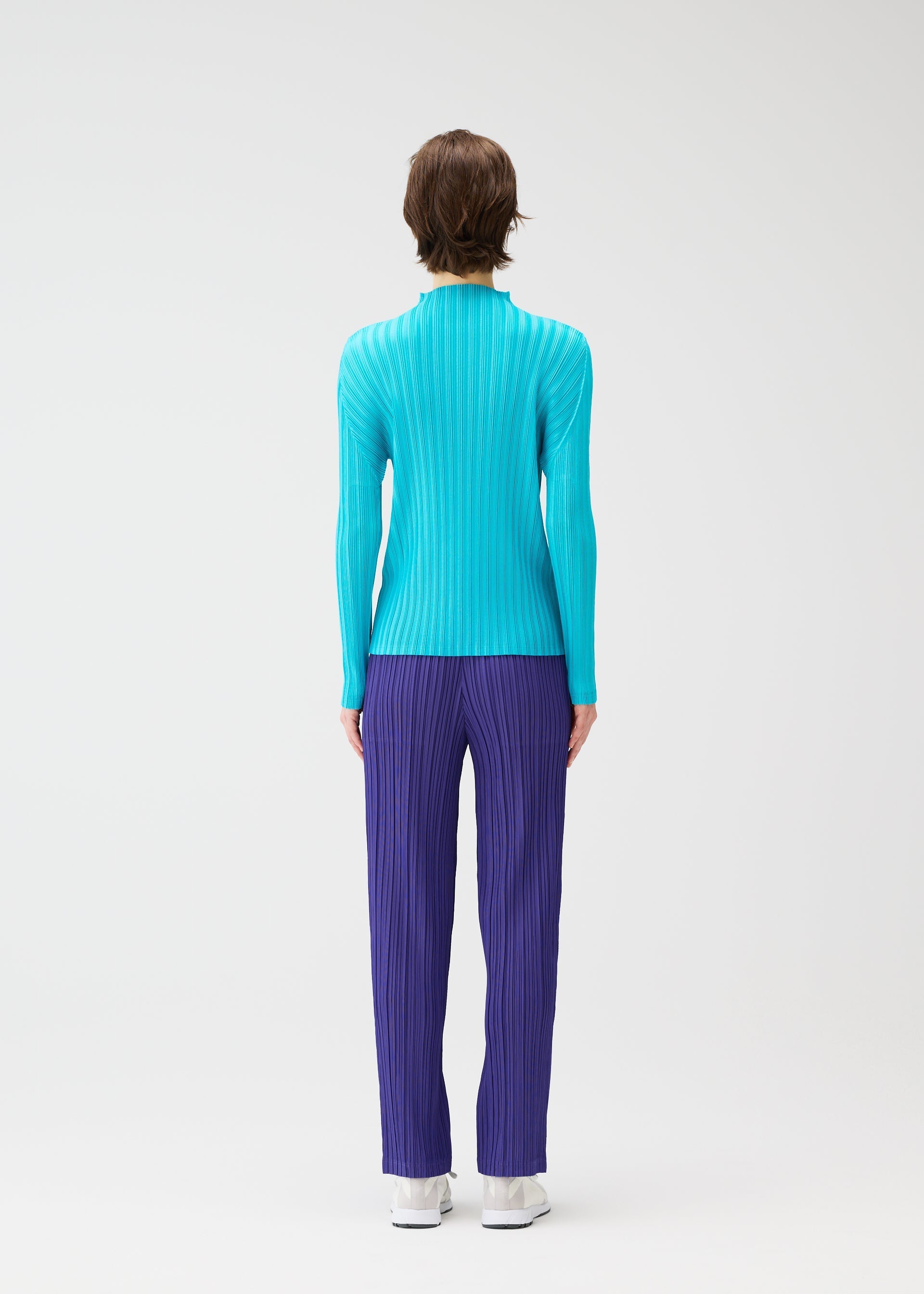 Zoomed view A person with short brown hair stands facing away, wearing the RIB PLEATS JANUARY set by PLEATS PLEASE ISSEY MIYAKE—a bright blue high-neck ribbed top and purple ribbed pants—paired with white shoes, against a plain white background.