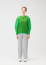 A woman smiles in the TATAMI APRIL long-sleeve top by PLEATS PLEASE ISSEY MIYAKE, featuring dolman sleeves and a red collar accent, paired with light gray pleated pants and white sneakers against a plain white background.