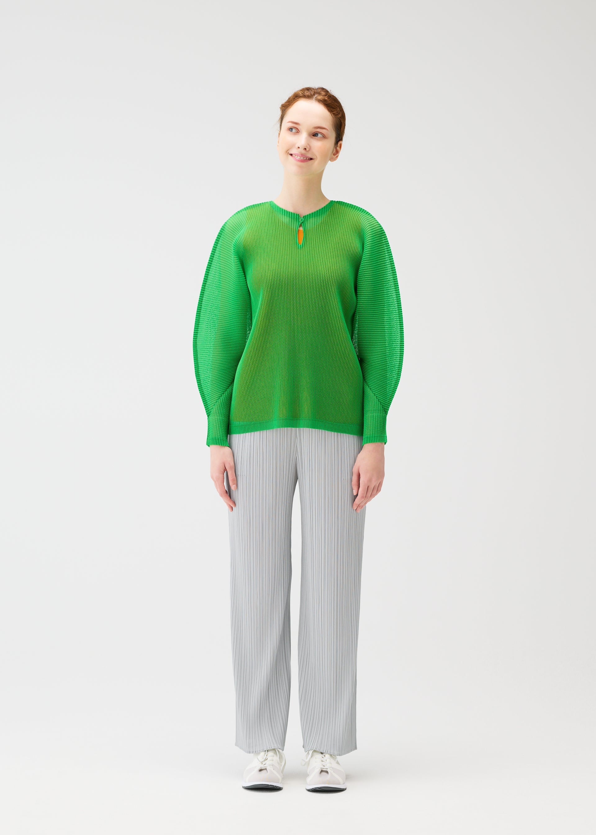 Zoomed view A woman smiles in the TATAMI APRIL long-sleeve top by PLEATS PLEASE ISSEY MIYAKE, featuring dolman sleeves and a red collar accent, paired with light gray pleated pants and white sneakers against a plain white background.