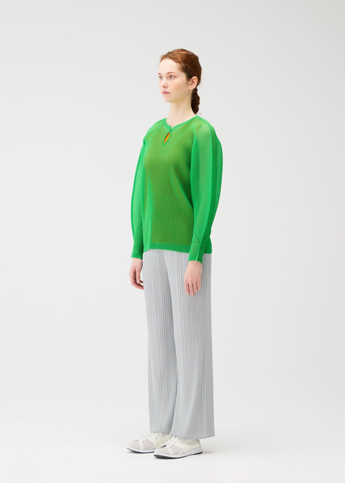 A person stands slightly to the side against a white background, wearing the TATAMI APRIL pleated wide-leg pants by PLEATS PLEASE ISSEY MIYAKE, styled with a sheer green mesh top and white sneakers. view 6 of 7