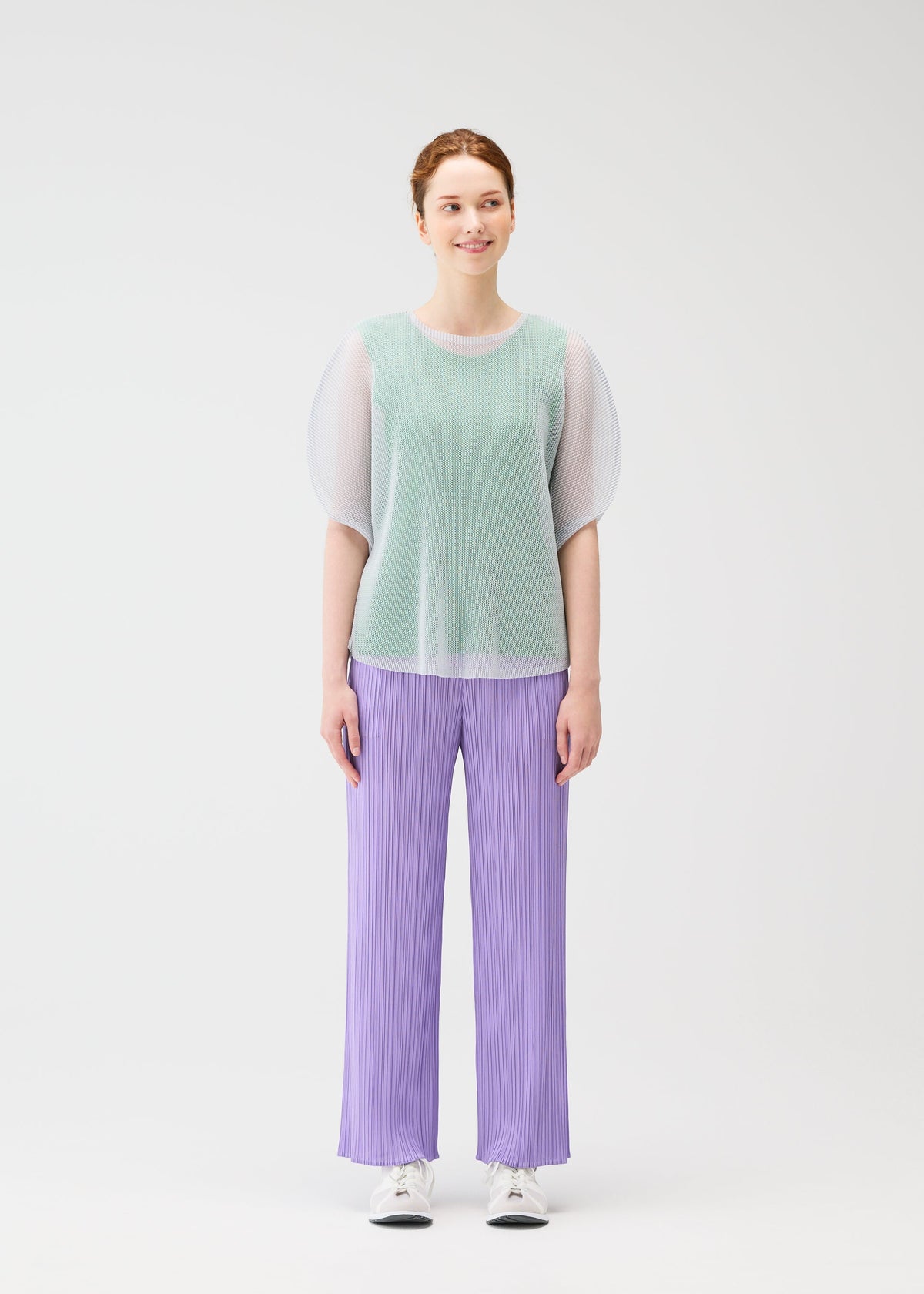 A woman with light skin and auburn hair stands against a white background, wearing PLEATS PLEASE ISSEY MIYAKE TATAMI APRIL mint green mesh top, lavender pleated pants, and white sneakers. view 6 of 8