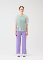 A woman with light skin and auburn hair stands against a white background, wearing PLEATS PLEASE ISSEY MIYAKE TATAMI APRIL mint green mesh top, lavender pleated pants, and white sneakers.