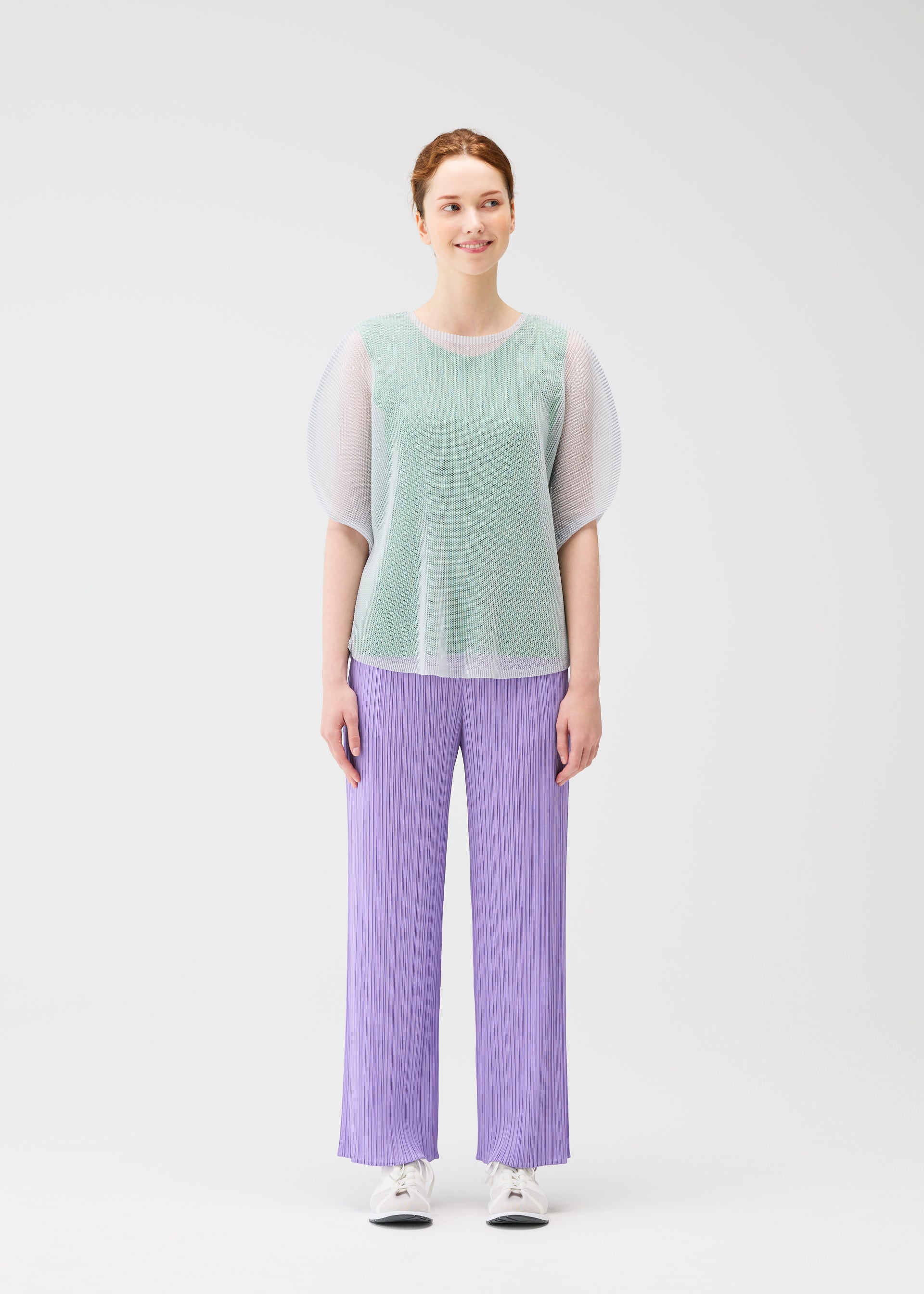 Zoomed view A woman with light skin and auburn hair stands against a white background, wearing PLEATS PLEASE ISSEY MIYAKE TATAMI APRIL mint green mesh top, lavender pleated pants, and white sneakers.
