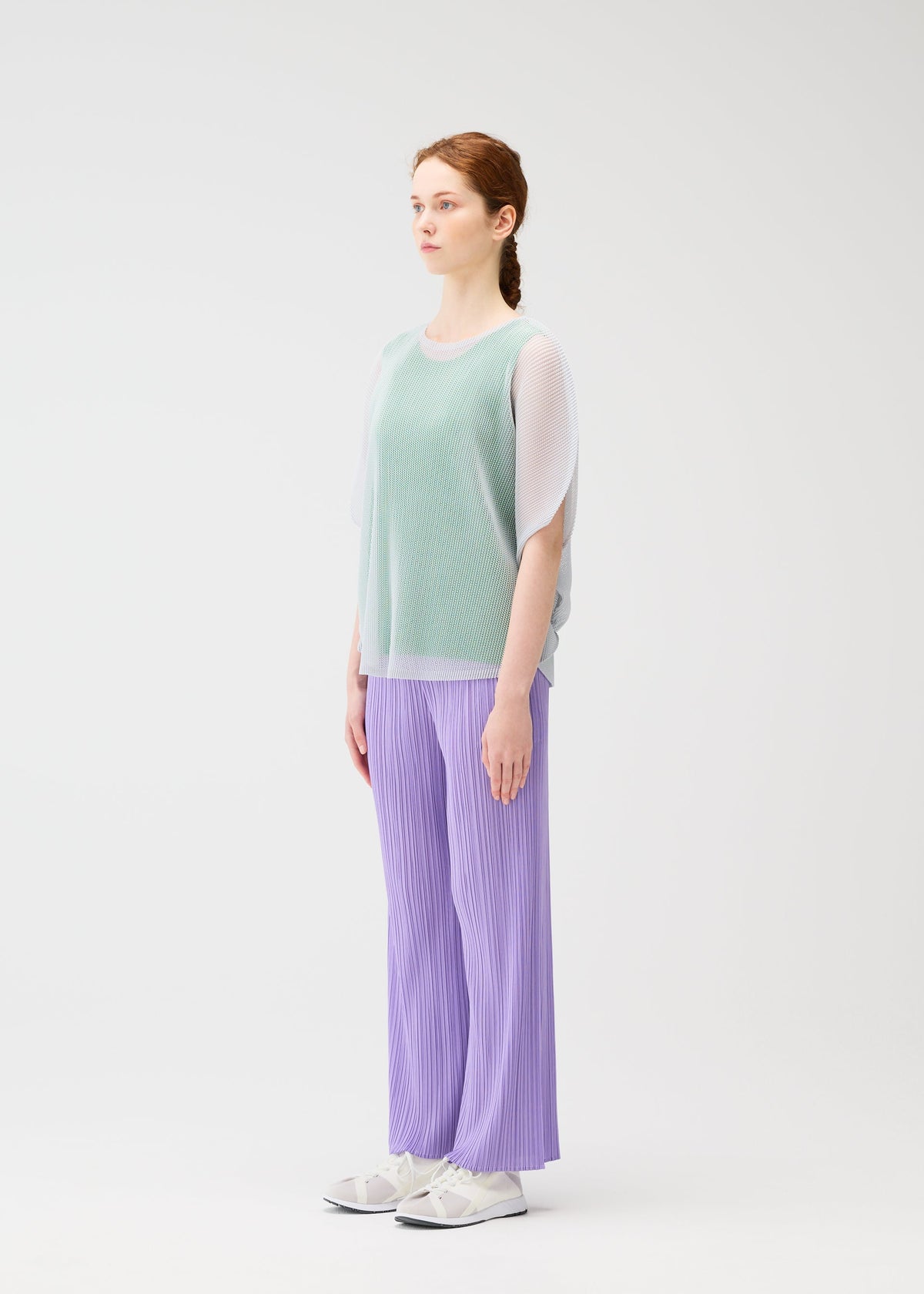 A light-skinned redhead with a braid stands sideways in the PLEATS PLEASE ISSEY MIYAKE TATAMI APRIL mint green mesh top over a white shirt, loose lavender pleated pants, and white sneakers against a plain white background. view 7 of 8