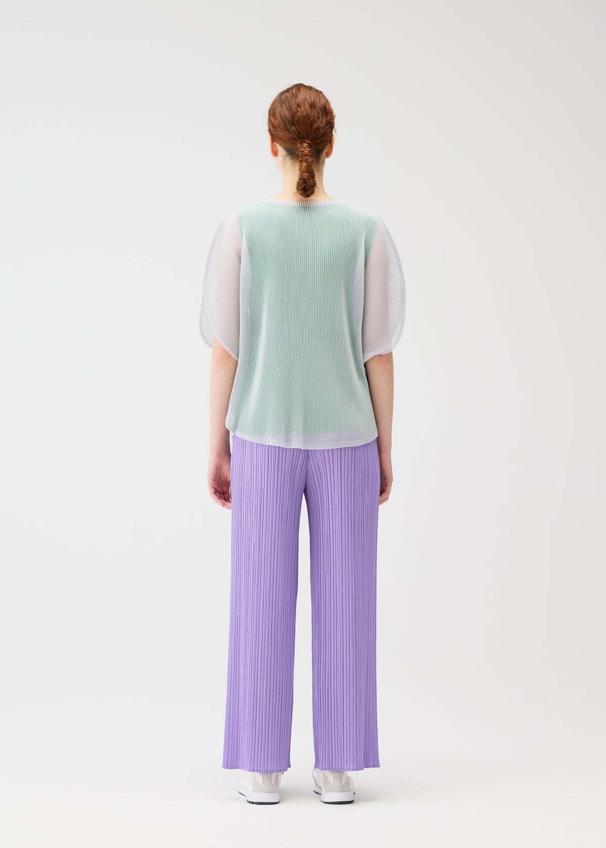A person with red hair in a low braid faces away, modeling the TATAMI APRIL by PLEATS PLEASE ISSEY MIYAKE: a sheer mint green mesh top, loose pleated lavender pants, and white shoes against a plain white background. view 8 of 8