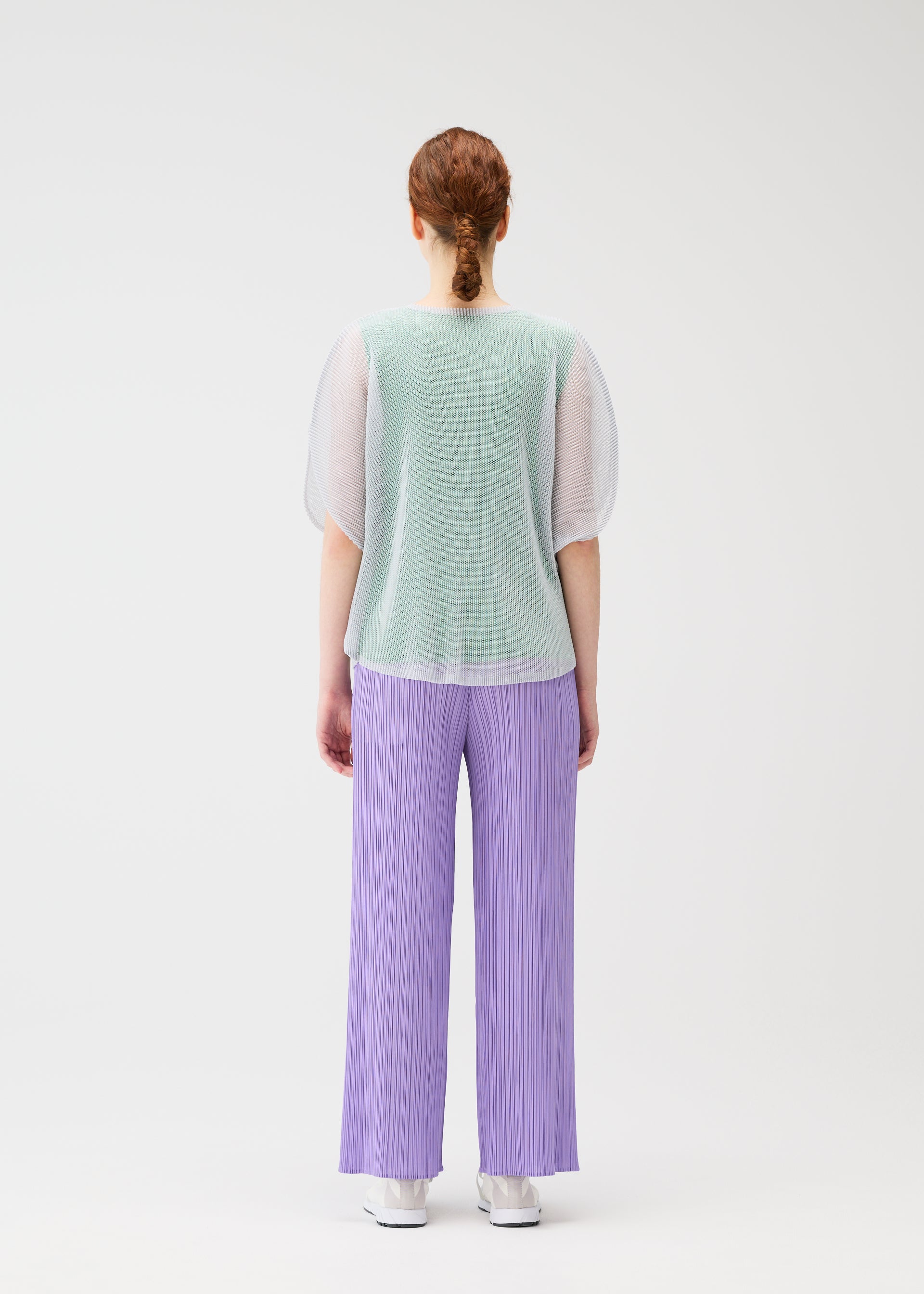 Zoomed view A person with red hair in a low braid faces away, modeling the TATAMI APRIL by PLEATS PLEASE ISSEY MIYAKE: a sheer mint green mesh top, loose pleated lavender pants, and white shoes against a plain white background.