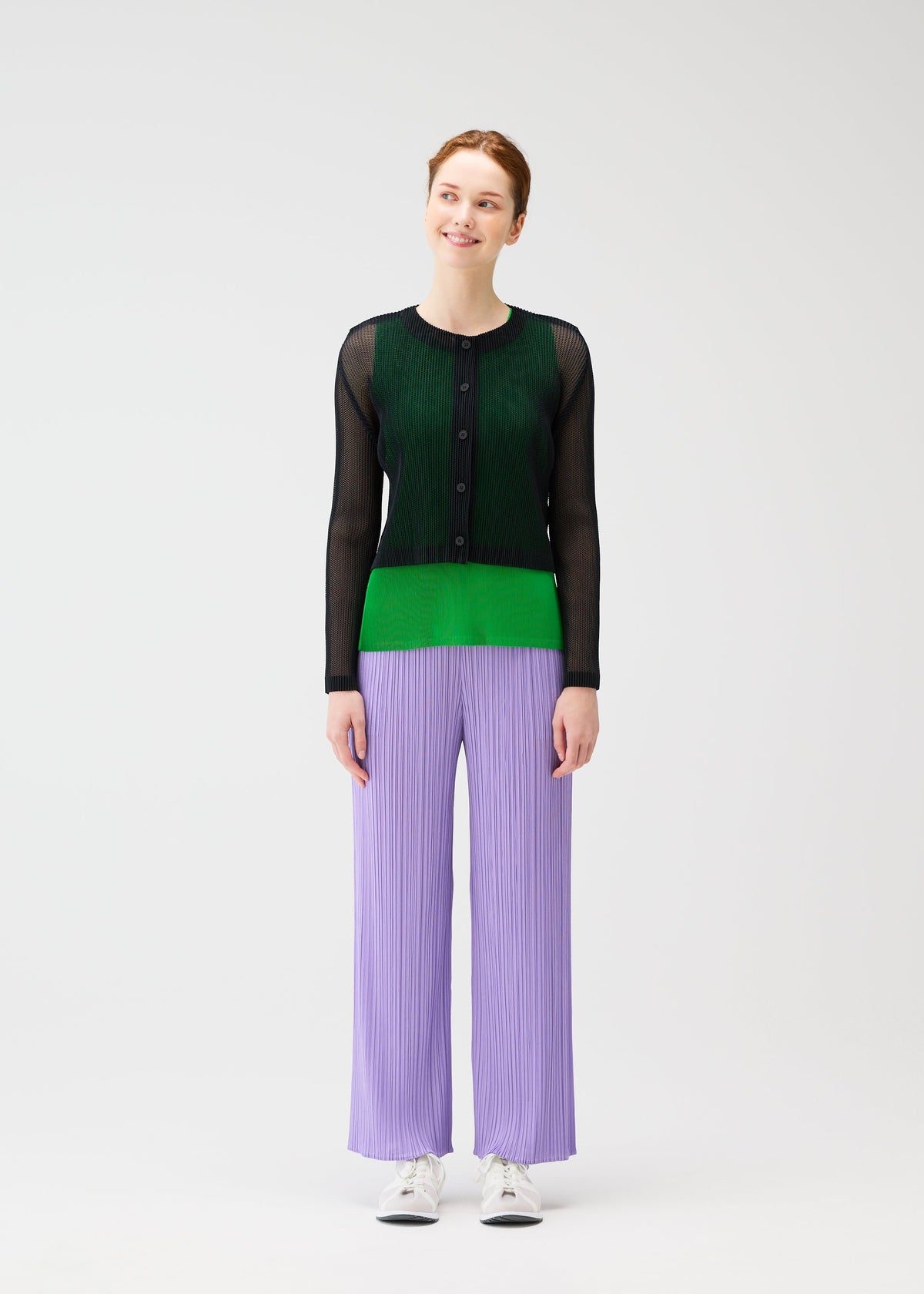 A person stands against a plain white background, smiling, wearing the TATAMI APRIL pleated pants by PLEATS PLEASE ISSEY MIYAKE in light purple, paired with a black and green mesh long-sleeve top and white sneakers. view 5 of 7