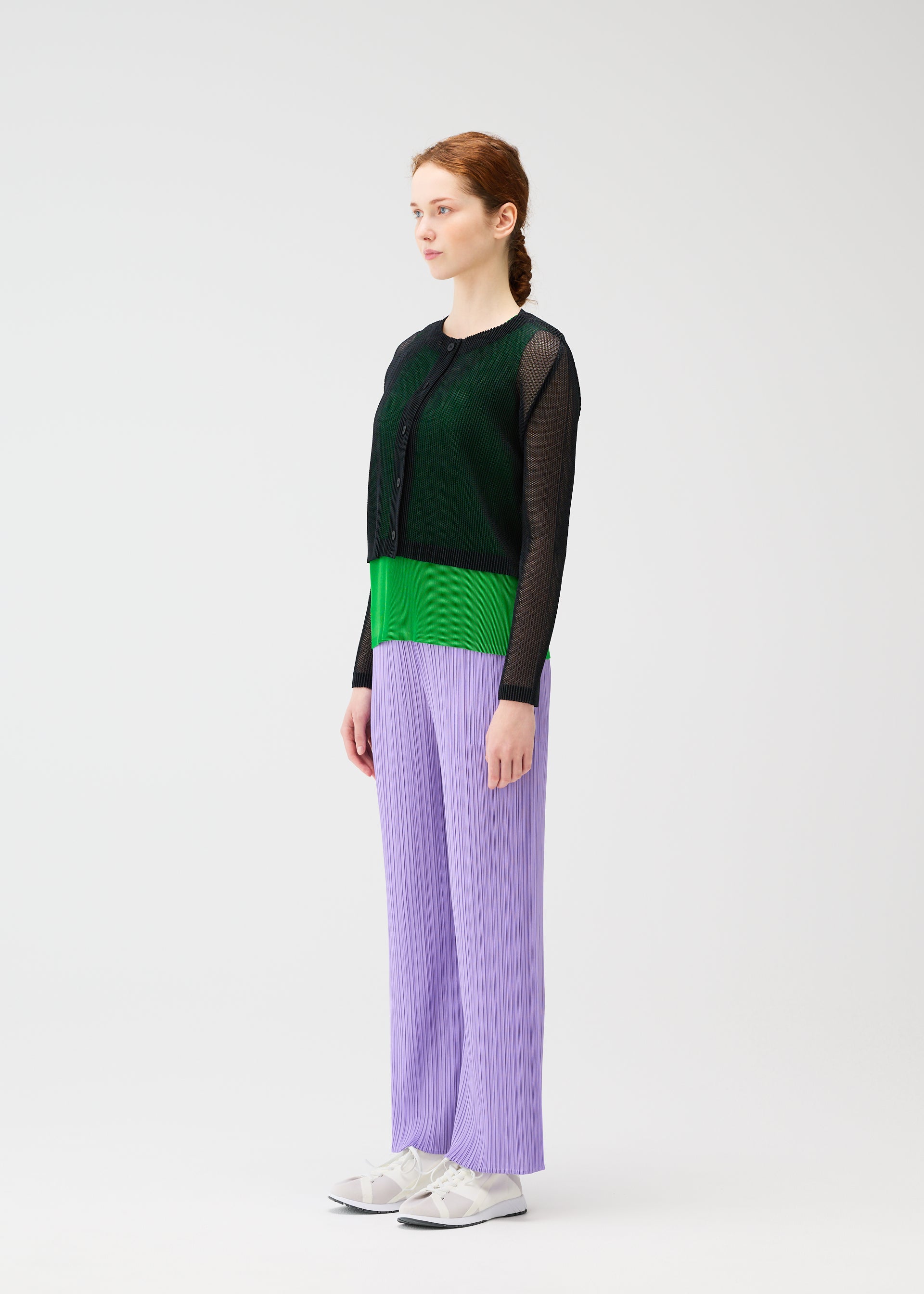 Zoomed view A woman with red hair in a braid stands sideways against a white background, wearing the TATAMI APRIL pleated lilac pants by PLEATS PLEASE ISSEY MIYAKE, paired with a sheer black mesh top over a green shirt and white sneakers.