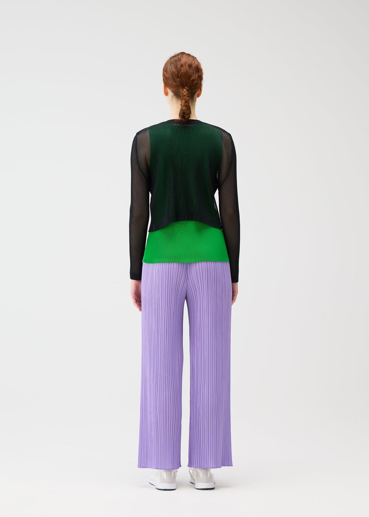 A person with red hair in a low braid faces away, wearing wide lilac TATAMI APRIL pants by PLEATS PLEASE ISSEY MIYAKE, black mesh long-sleeve top, green shirt, and white sneakers against a plain white background. view 7 of 7