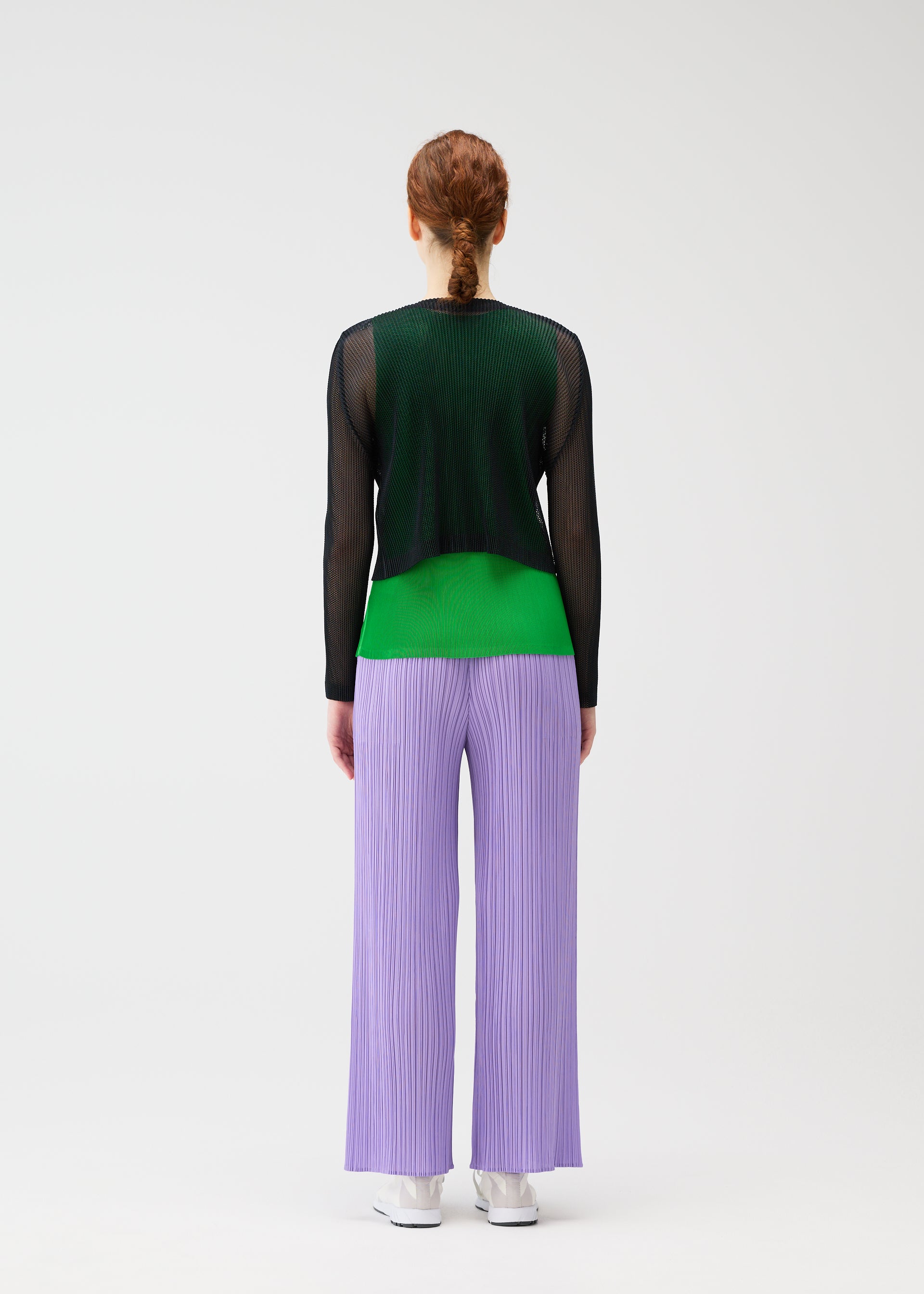 Zoomed view A person with red hair in a low braid faces away, wearing wide lilac TATAMI APRIL pants by PLEATS PLEASE ISSEY MIYAKE, black mesh long-sleeve top, green shirt, and white sneakers against a plain white background.