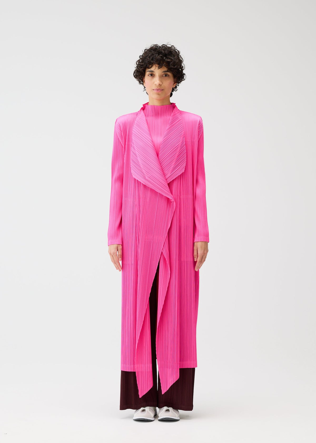 A person with short curly hair models the MONTHLY COLORS : JANUARY bright pink buttoned long coat by PLEATS PLEASE ISSEY MIYAKE, styled over dark pants and white shoes, standing against a plain white background. view 7 of 10
