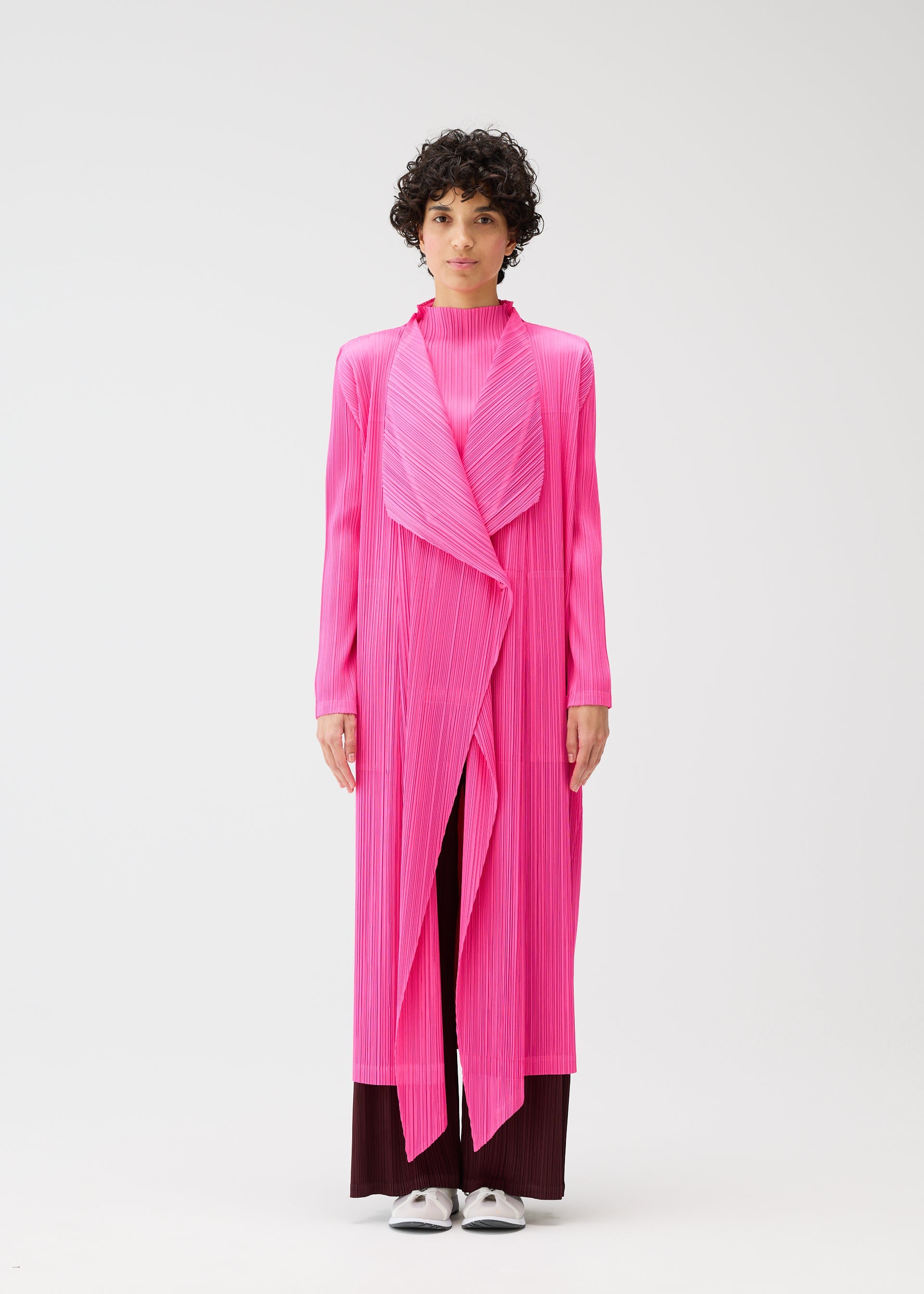 Zoomed view A person with short curly hair models the MONTHLY COLORS : JANUARY bright pink buttoned long coat by PLEATS PLEASE ISSEY MIYAKE, styled over dark pants and white shoes, standing against a plain white background.