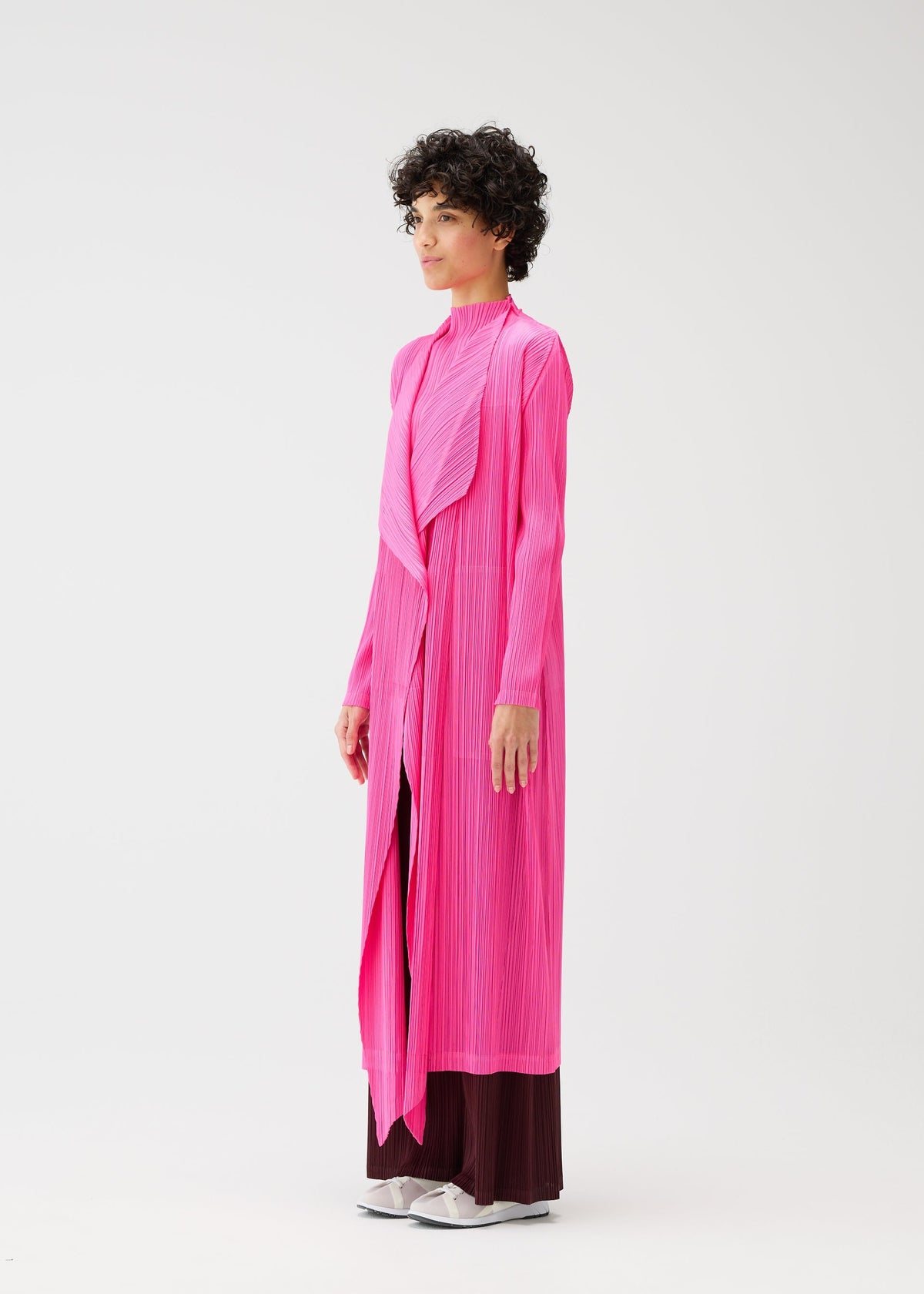 A person stands sideways against a plain white background, wearing the bright pink MONTHLY COLORS : JANUARY long pleated coat from PLEATS PLEASE ISSEY MIYAKE over dark wide-leg pants and white shoes. view 8 of 10