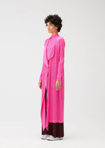 A person stands sideways against a plain white background, wearing the bright pink MONTHLY COLORS : JANUARY long pleated coat from PLEATS PLEASE ISSEY MIYAKE over dark wide-leg pants and white shoes.