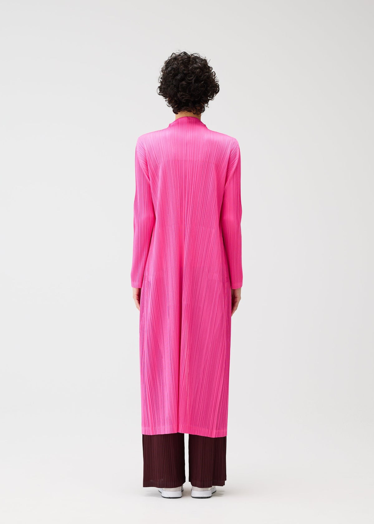 A person with short curly hair stands facing away, wearing the MONTHLY COLORS : JANUARY bright pink pleated coat by PLEATS PLEASE ISSEY MIYAKE over dark brown wide-leg pants and white shoes, against a plain white background. view 9 of 10