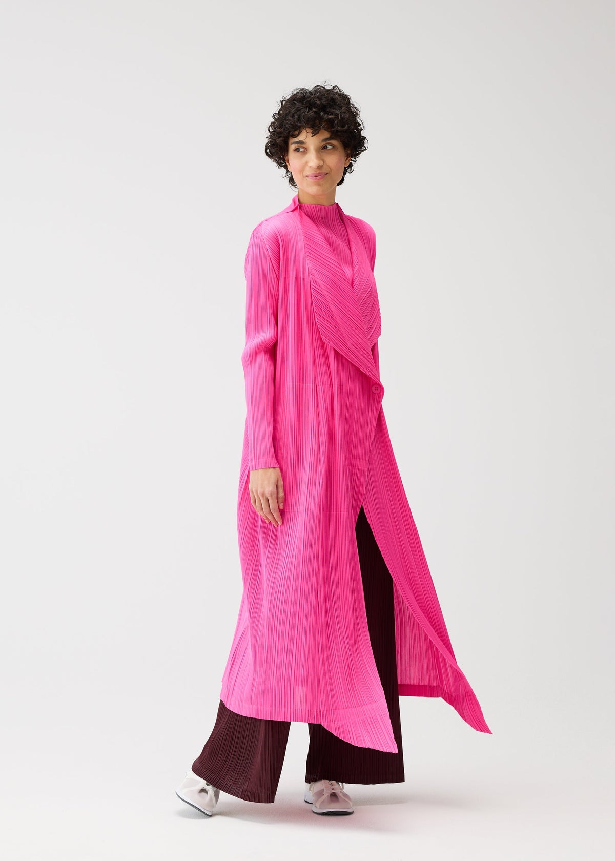 A person with short curly hair wears the MONTHLY COLORS : JANUARY bright pink, buttoned long coat by PLEATS PLEASE ISSEY MIYAKE over burgundy wide-leg pants and white shoes, walking against a plain light background. view 10 of 10
