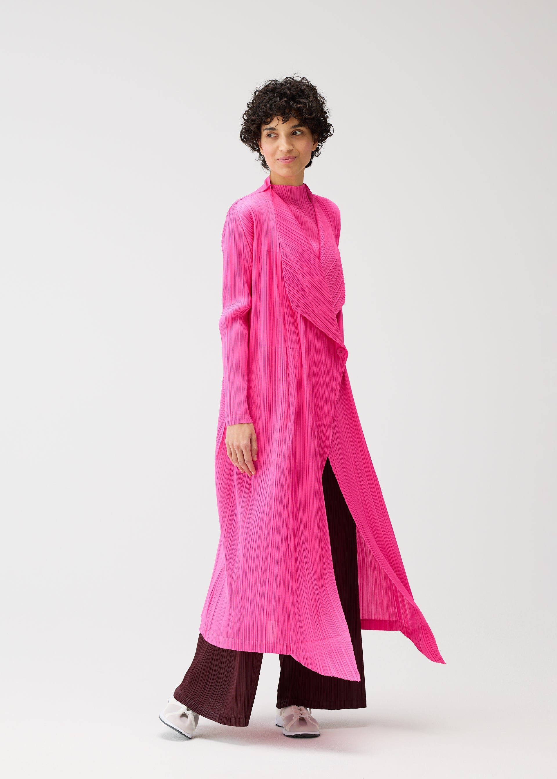 Zoomed view A person with short curly hair wears the MONTHLY COLORS : JANUARY bright pink, buttoned long coat by PLEATS PLEASE ISSEY MIYAKE over burgundy wide-leg pants and white shoes, walking against a plain light background.