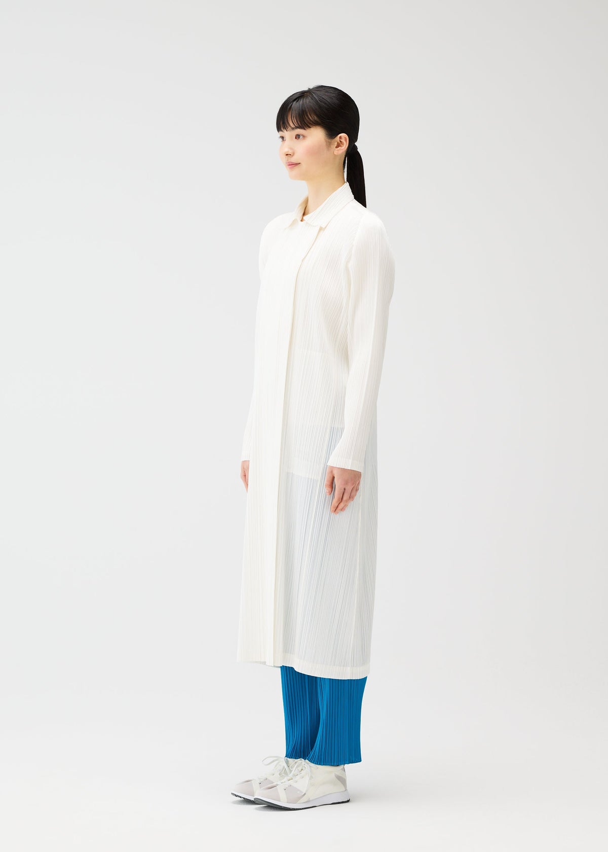 A woman in profile poses against a plain white backdrop, wearing PLEATS PLEASE ISSEY MIYAKE's MONTHLY COLORS : FEBRUARY— a straight silhouette tunic with bright blue pleated pants and white sneakers, styled with sleek black hair in a low ponytail. view 9 of 12