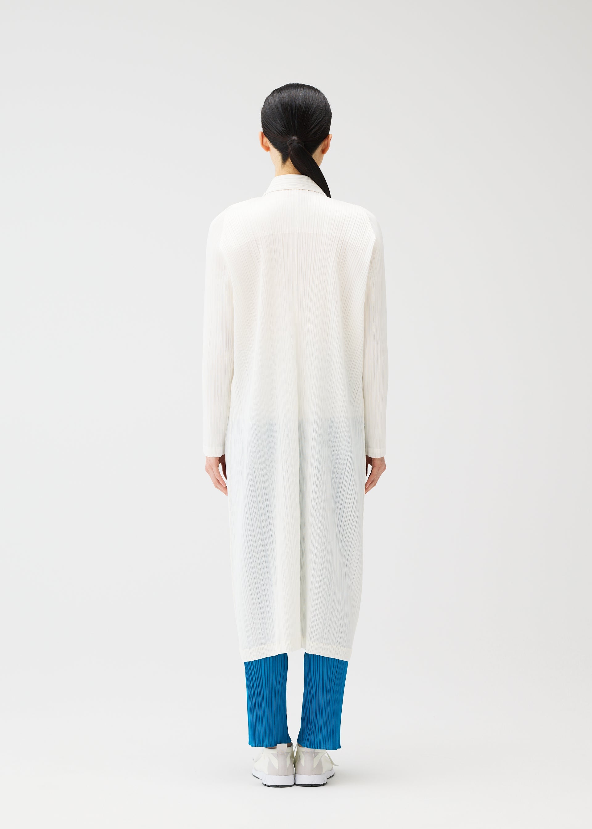 Zoomed view A person with long dark hair in a low ponytail stands facing away, wearing the PLEATS PLEASE ISSEY MIYAKE MONTHLY COLORS : FEBRUARY straight silhouette collared long coat over bright blue pleated pants and white shoes, against a plain white background.