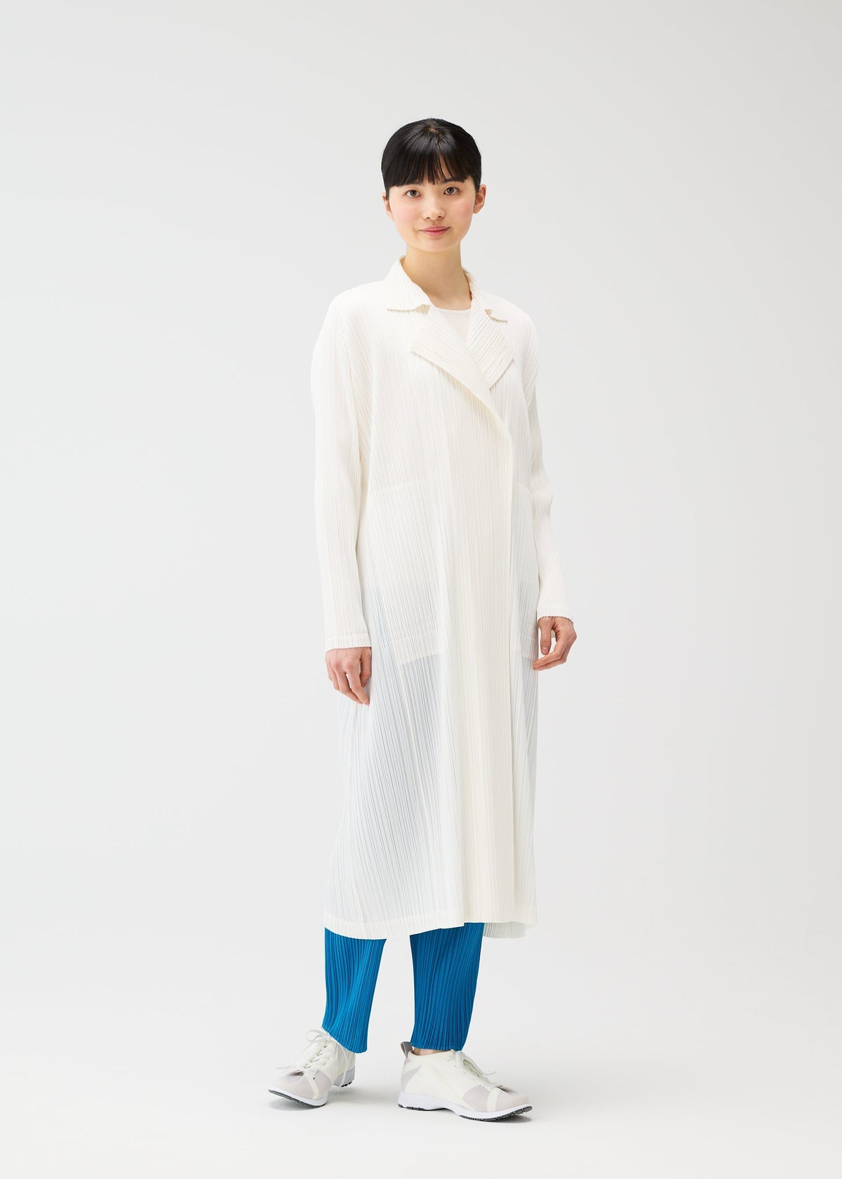 Against a plain white background, a person with short black hair and a relaxed expression wears PLEATS PLEASE ISSEY MIYAKE’s MONTHLY COLORS : FEBRUARY—a collared long coat over a white top, bright blue pleated pants, and gray sneakers. view 12 of 12