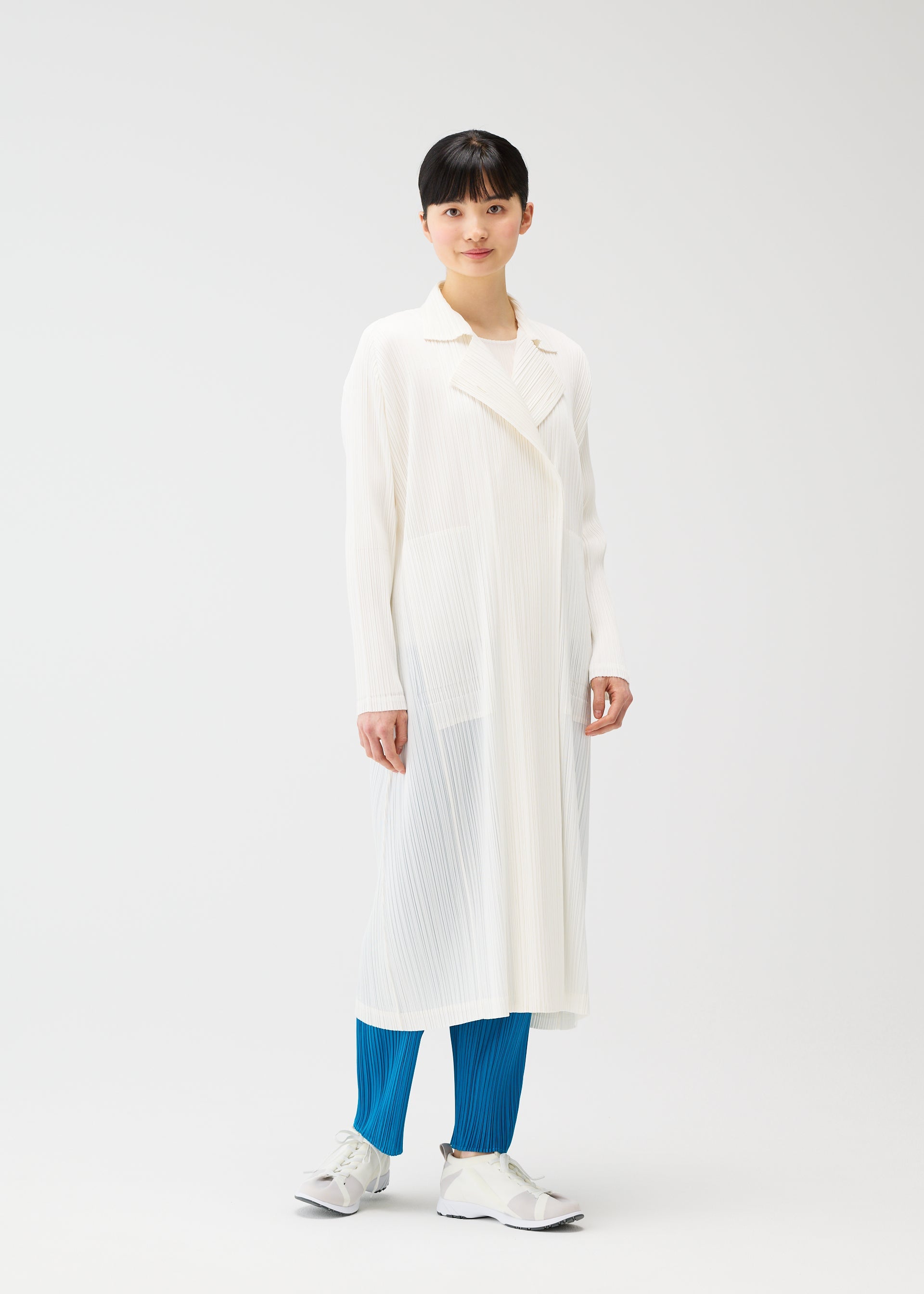Zoomed view Against a plain white background, a person with short black hair and a relaxed expression wears PLEATS PLEASE ISSEY MIYAKE’s MONTHLY COLORS : FEBRUARY—a collared long coat over a white top, bright blue pleated pants, and gray sneakers.