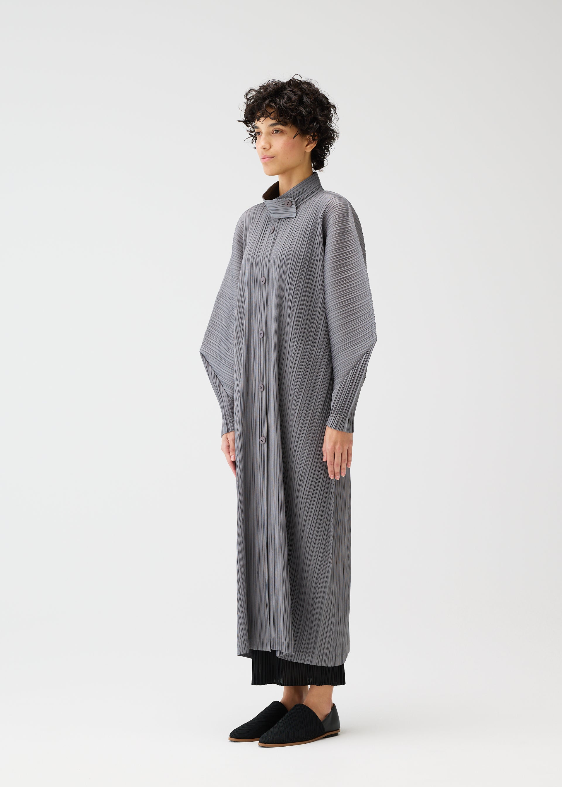 Zoomed view A person with short, curly hair stands against a white background wearing the SKYLINE long buttoned coat with dolman sleeves and a standing collar from PLEATS PLEASE ISSEY MIYAKE, paired with black pants and slip-on shoes.