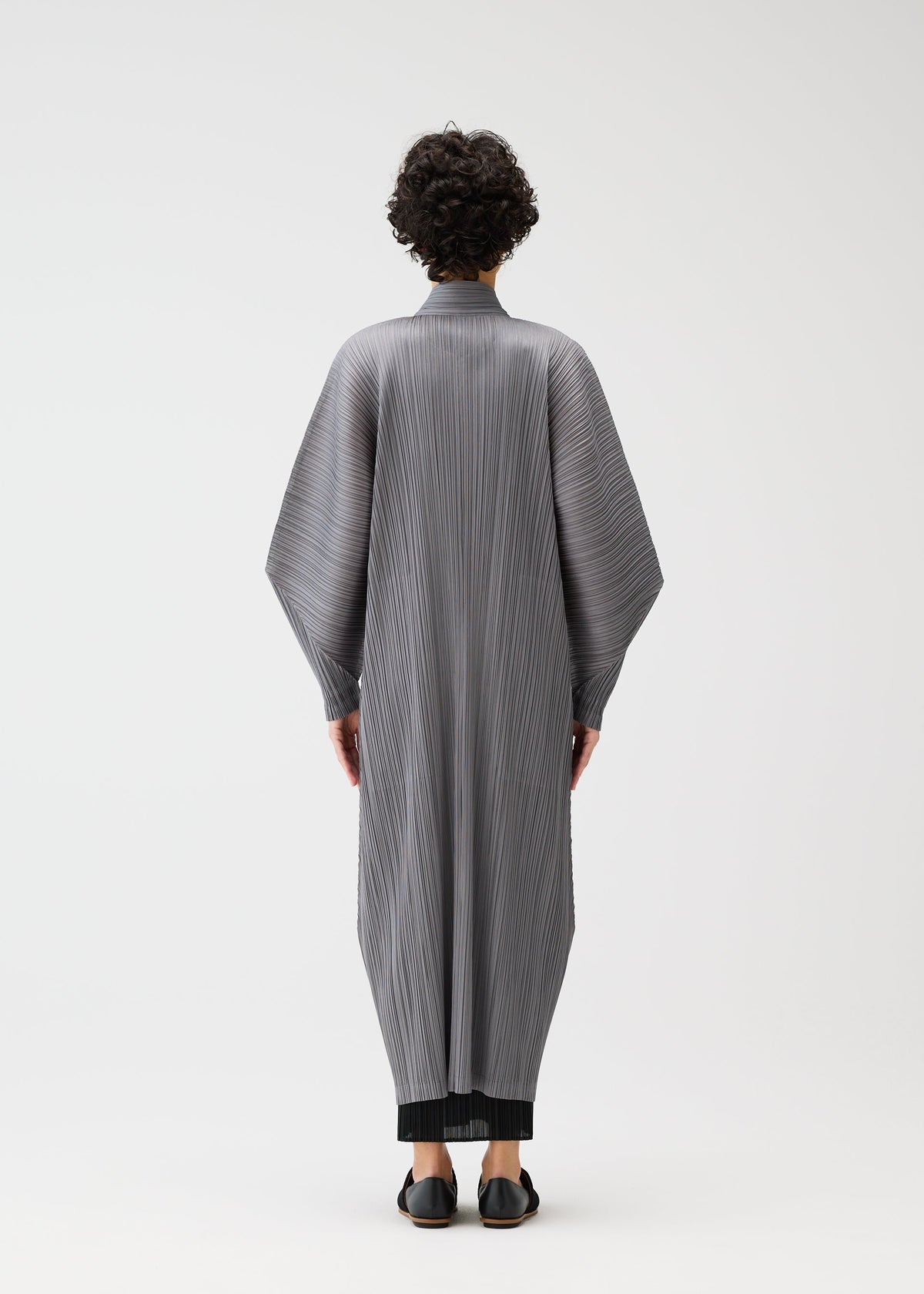 A person with short curly hair stands facing away, modeling the SKYLINE long buttoned coat with wide dolman sleeves and standing collar by PLEATS PLEASE ISSEY MIYAKE, over black pants and shoes, set against a plain white background. view 8 of 9