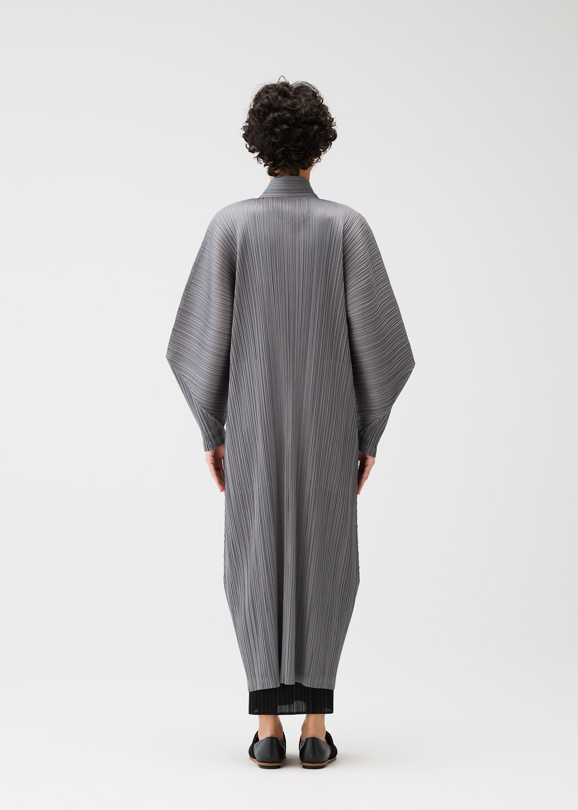 Zoomed view A person with short curly hair stands facing away, modeling the SKYLINE long buttoned coat with wide dolman sleeves and standing collar by PLEATS PLEASE ISSEY MIYAKE, over black pants and shoes, set against a plain white background.