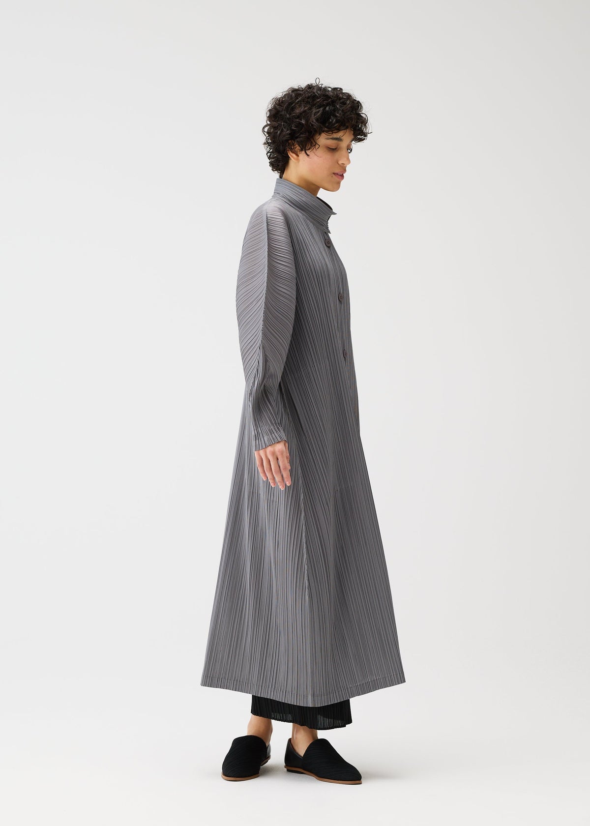 A person with short curly hair stands in profile, wearing the SKYLINE long buttoned coat-dress by PLEATS PLEASE ISSEY MIYAKE over black pants and black slip-on shoes, set against a plain white background. view 9 of 9