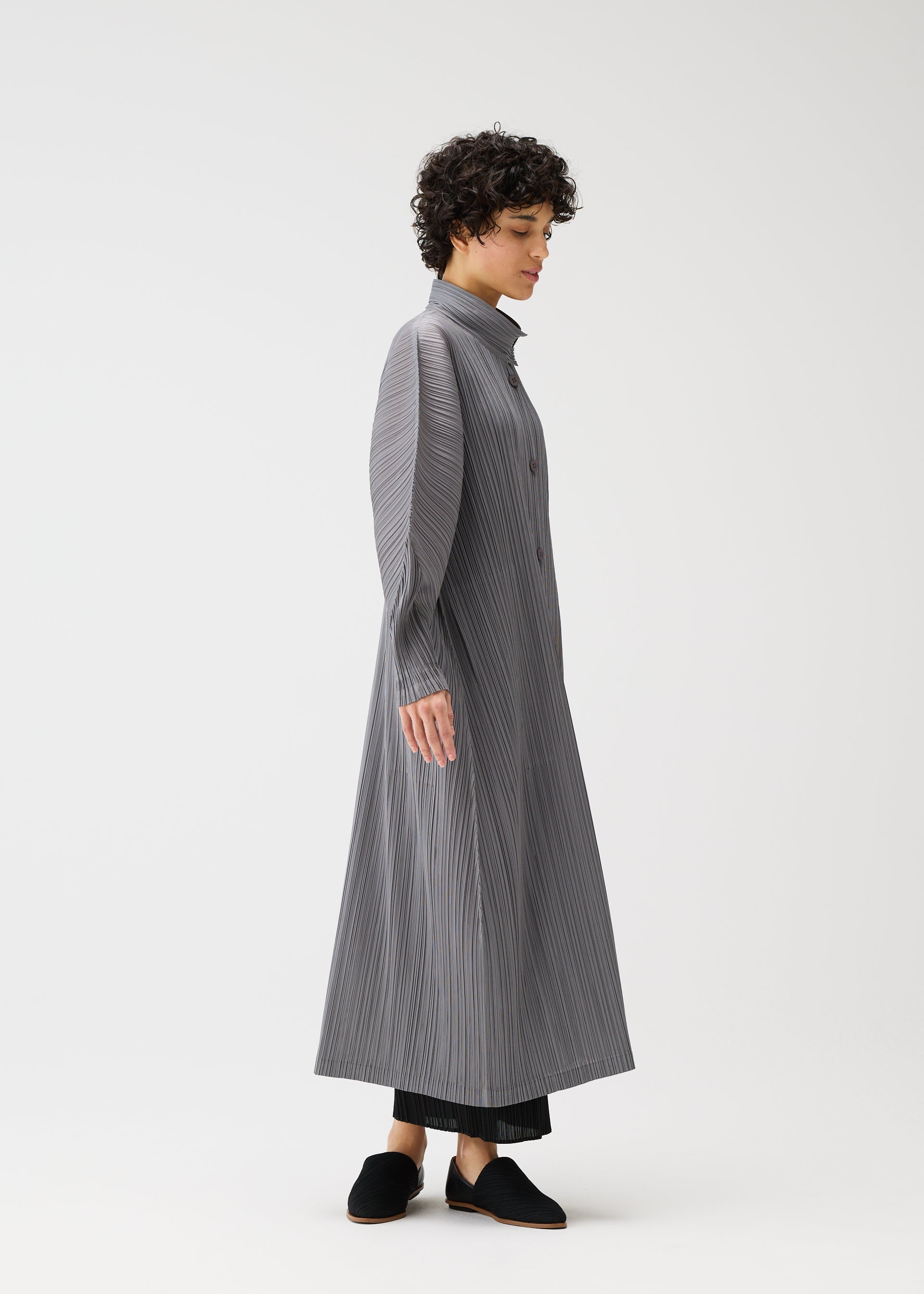Zoomed view A person with short curly hair stands in profile, wearing the SKYLINE long buttoned coat-dress by PLEATS PLEASE ISSEY MIYAKE over black pants and black slip-on shoes, set against a plain white background.